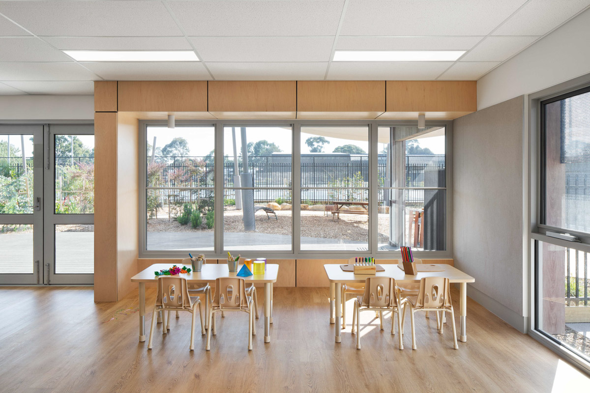 A bright kindergarten classroom with timber tables and chairs arranged near large windows. Colourful craft materials and pencils are on the tables, and the outdoor play area is visible through the glass doors.