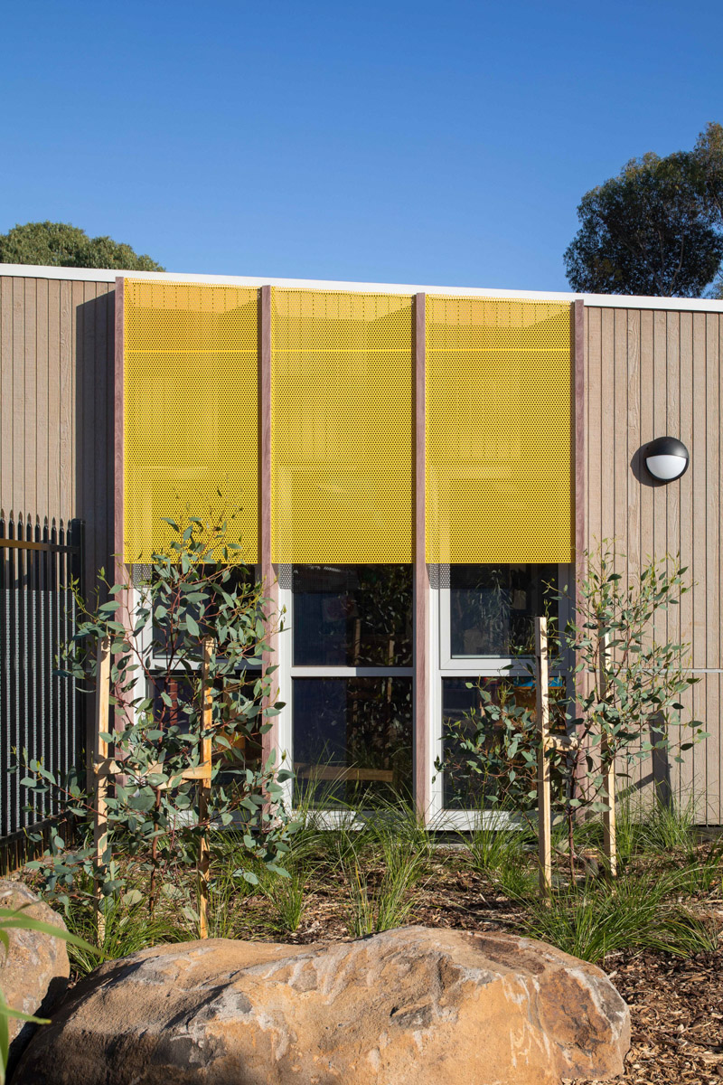 The exterior of the kindergarten with vertical timber panels and bright yellow perforated screens above the windows. There are young native plants and large rocks in the landscaped garden bed.