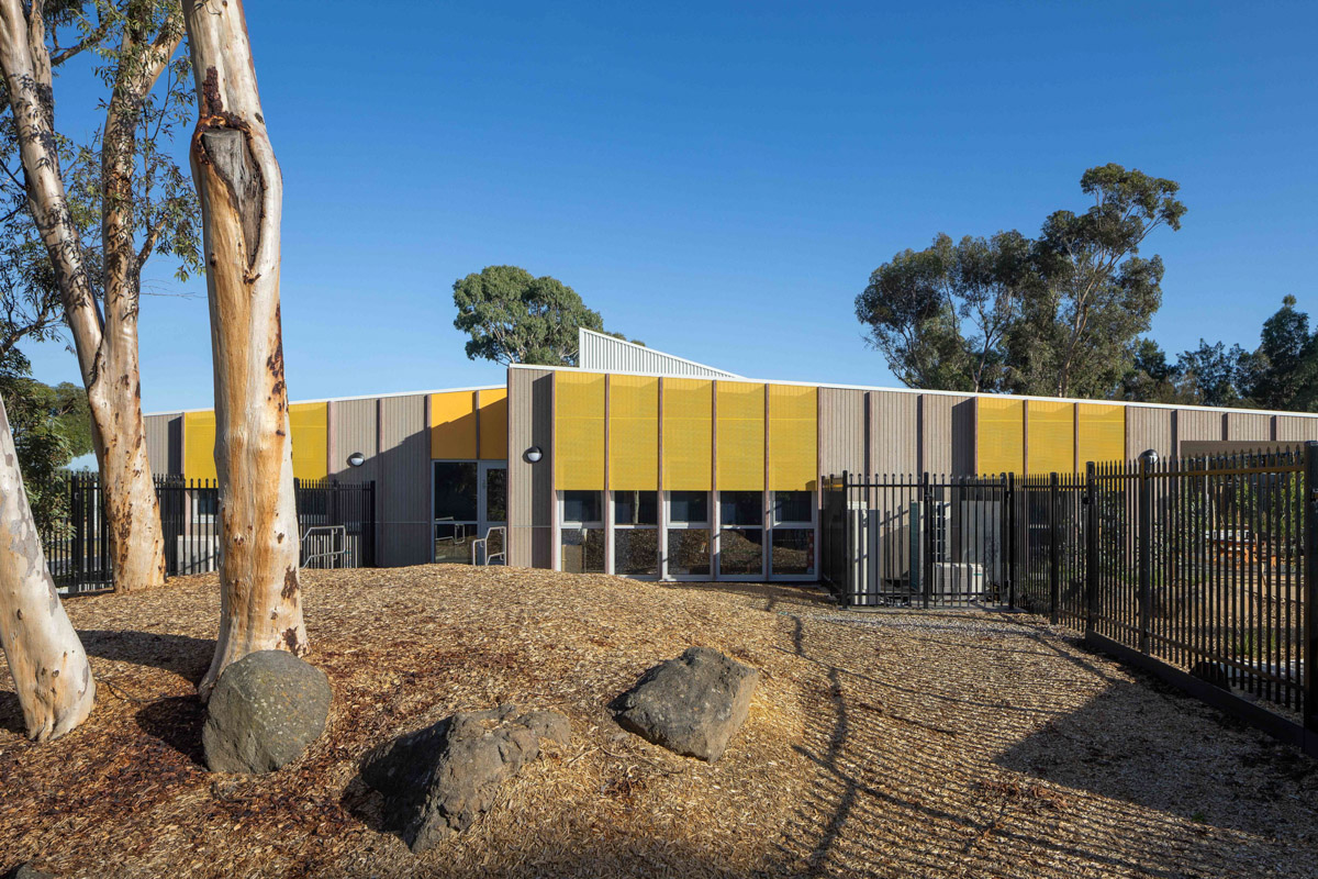 The exterior of the kindergarten viewed from the garden, with timber walls and yellow perforated screens. A black metal fence runs along the perimeter and gum trees stand in the foreground.