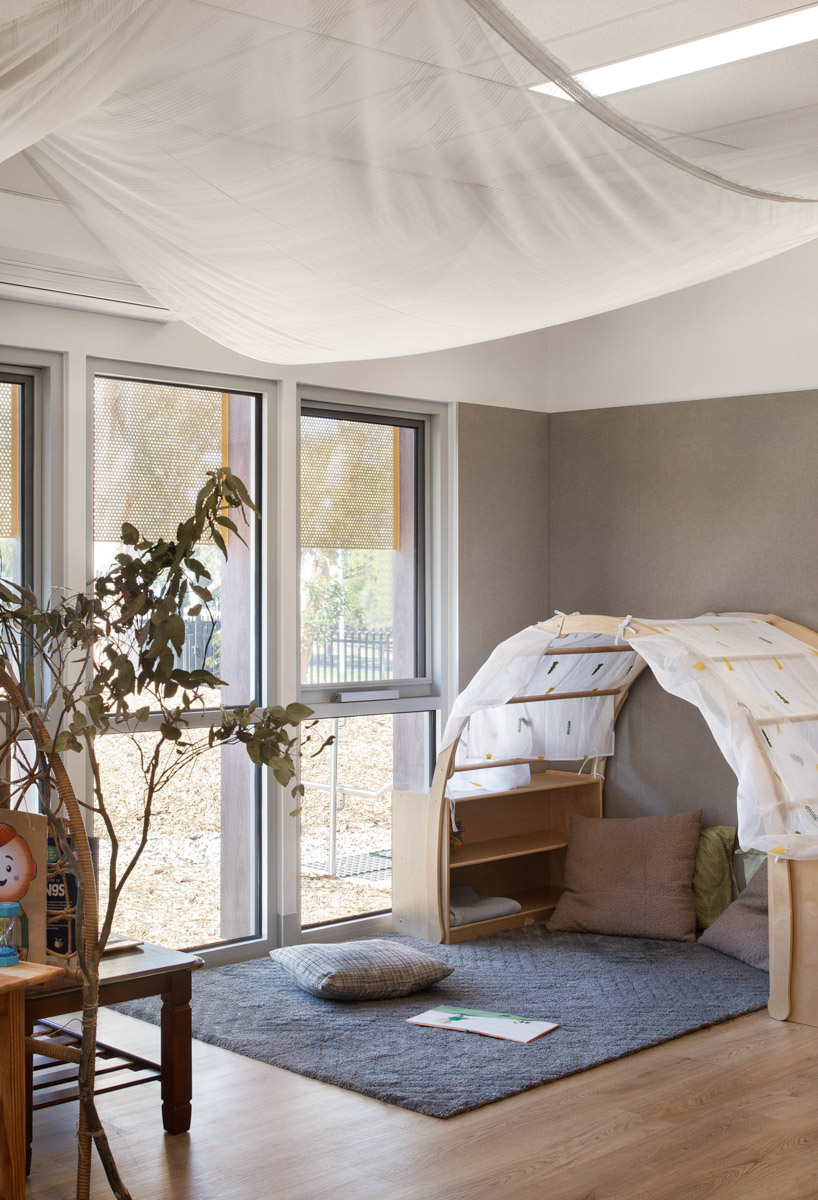 A cosy reading corner in the kindergarten with a small timber canopy structure draped in white fabric. Cushions and a rug are on the floor, and natural light streams through large windows with perforated screens.
