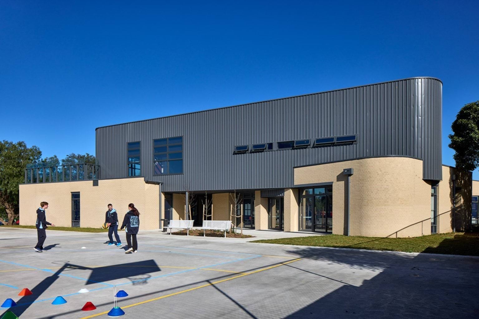 The exterior of a two-storey school building with curved grey cladding and beige brick walls. Students are playing on a paved area marked with sports lines and cones.