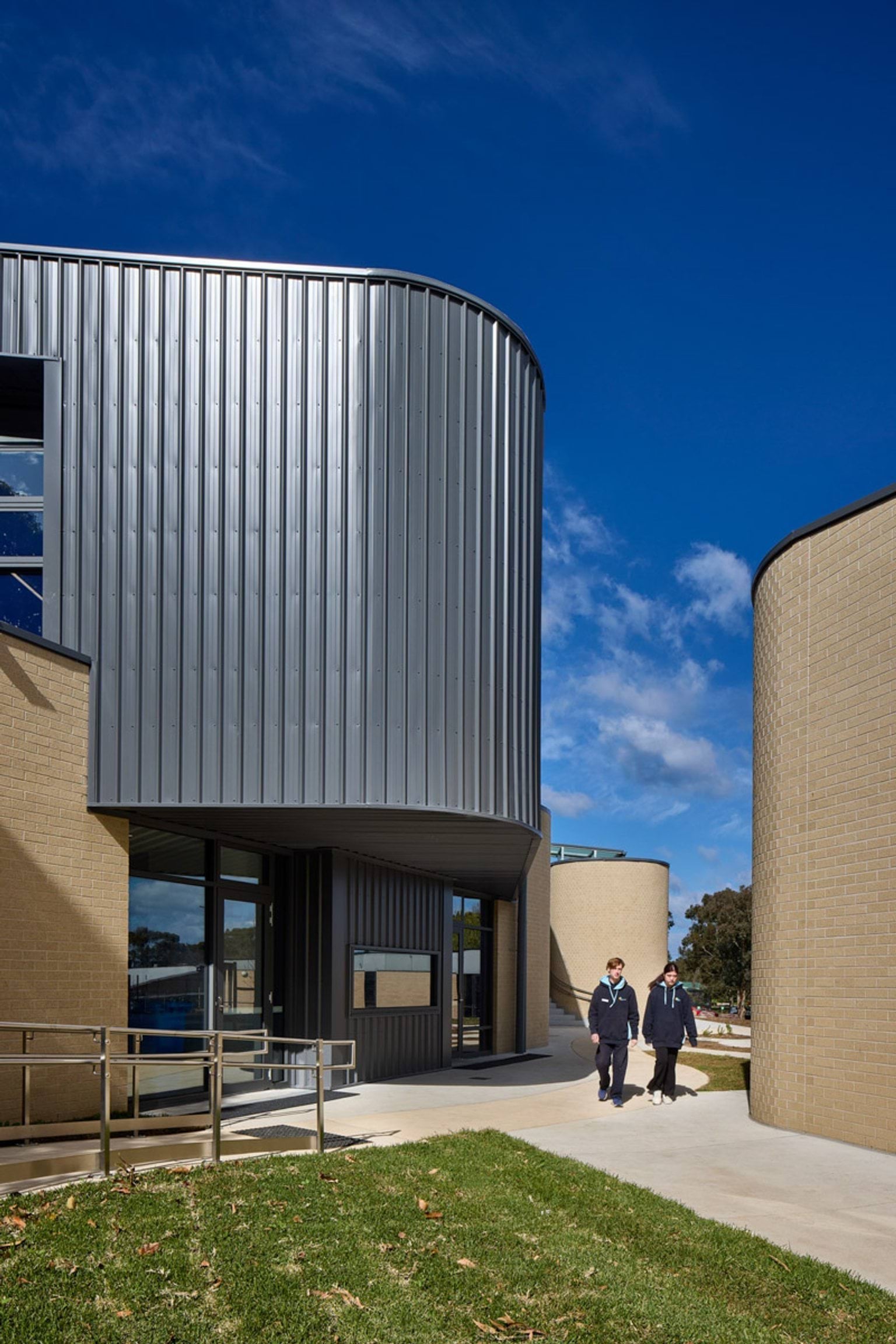 The school entrance with curved grey metal exterior and beige brick walls. Two students walk along a concrete path bordered by grass under a clear blue sky.