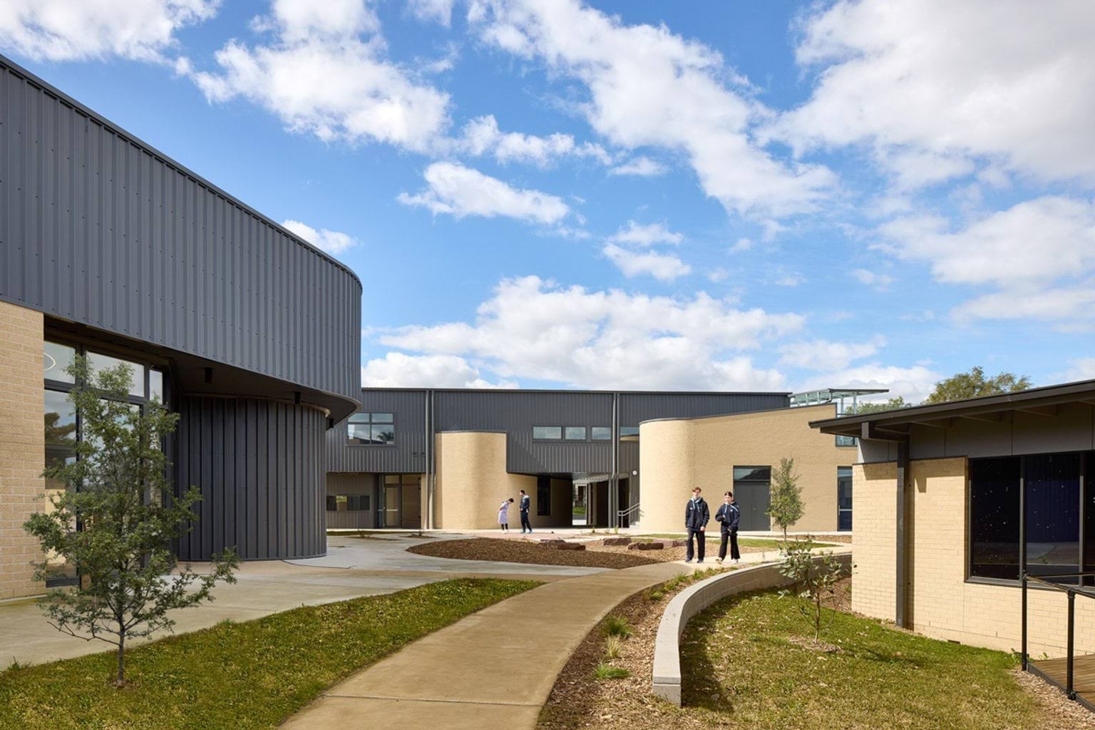 A landscaped courtyard with curved concrete paths and garden beds. Students are walking and talking near the modern school building.