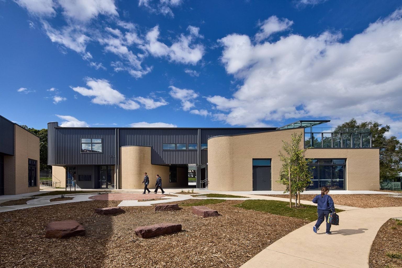 The exterior of the school with curved beige brick walls and grey cladding. Students walk along landscaped paths with mulch and stone features.