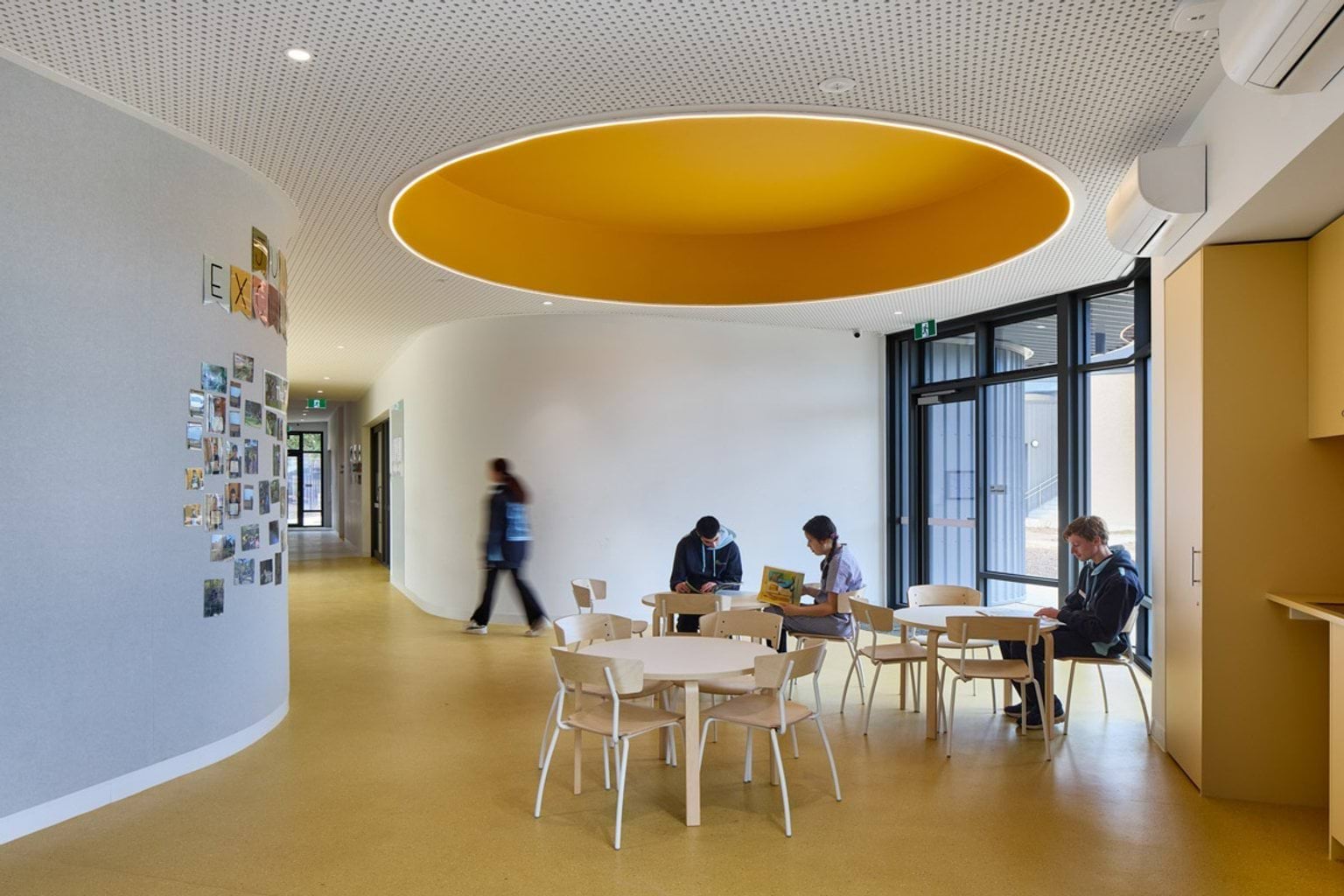 An open learning area with round tables and chairs beneath a large circular ceiling recess painted yellow. A wall displays photos and the space is well-lit by natural light.