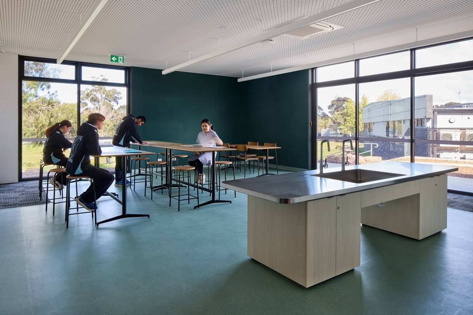 A classroom with green flooring, large windows, and high benches arranged in rows. A central island bench with a sink is in the foreground.