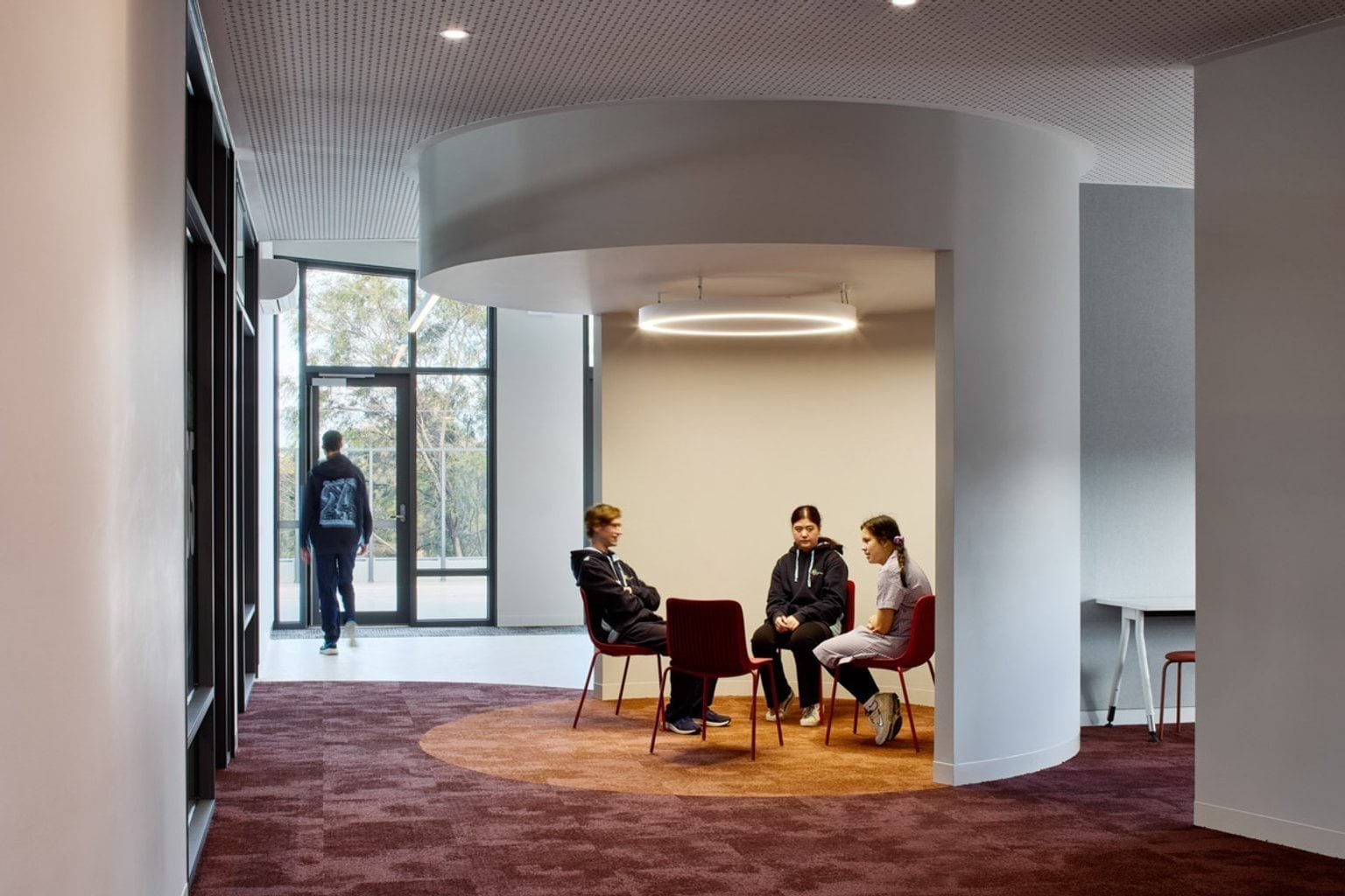 An interior space with a circular ceiling recess and pendant light above a group of chairs. A student walks toward glass doors in the background.