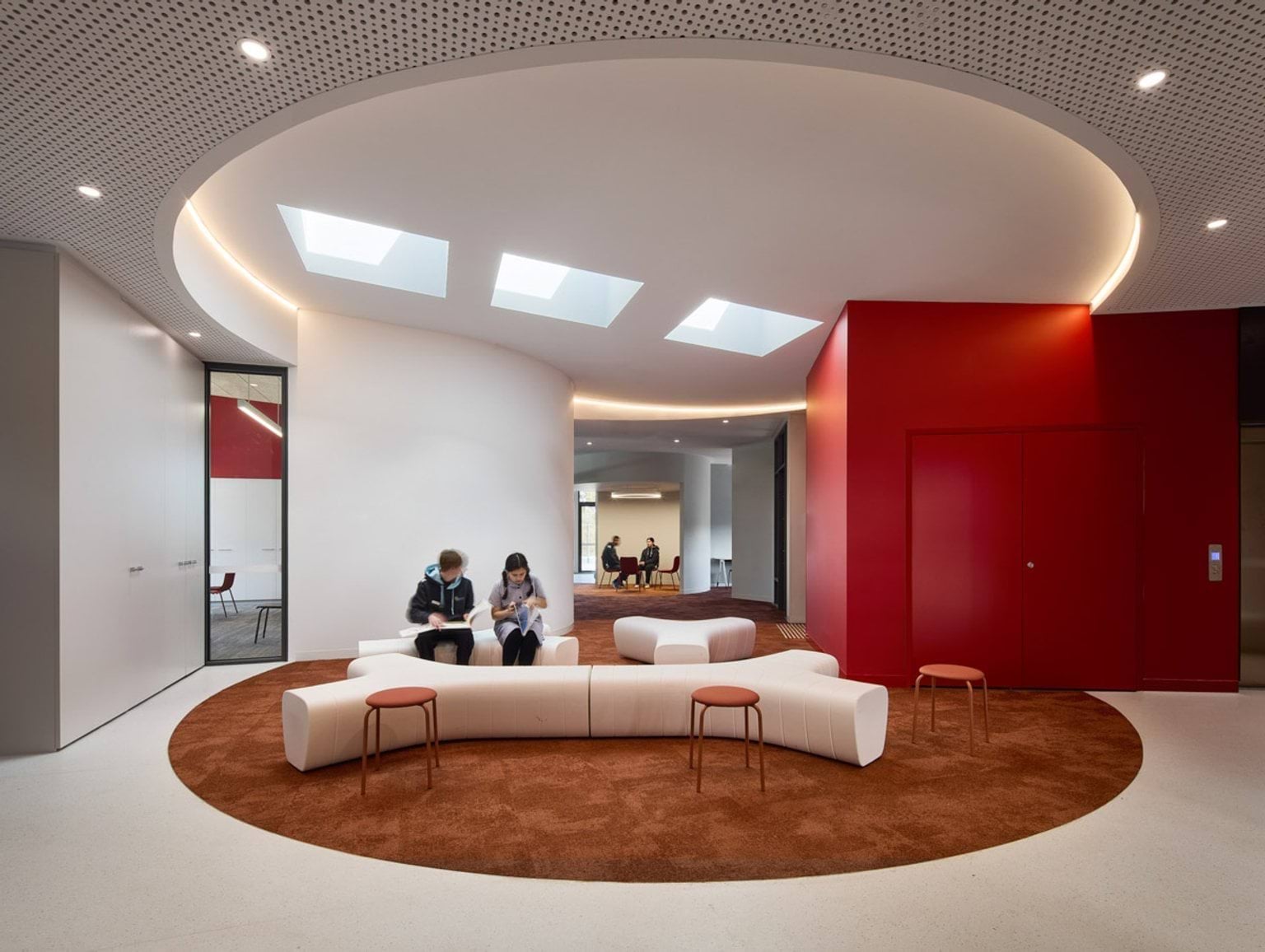 A large open lounge with curved walls, skylights, and a circular ceiling recess. White modular seating and red accents create a modern look.