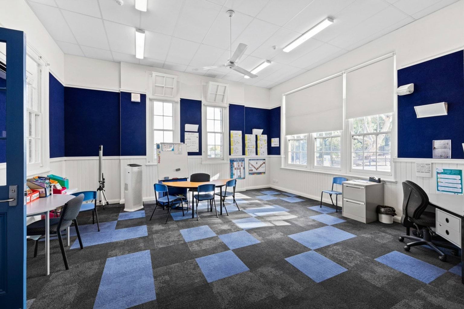 A classroom with blue and grey carpet tiles, white walls, and large windows. Tables and chairs are arranged for small group work, and teaching materials are displayed on the walls.
