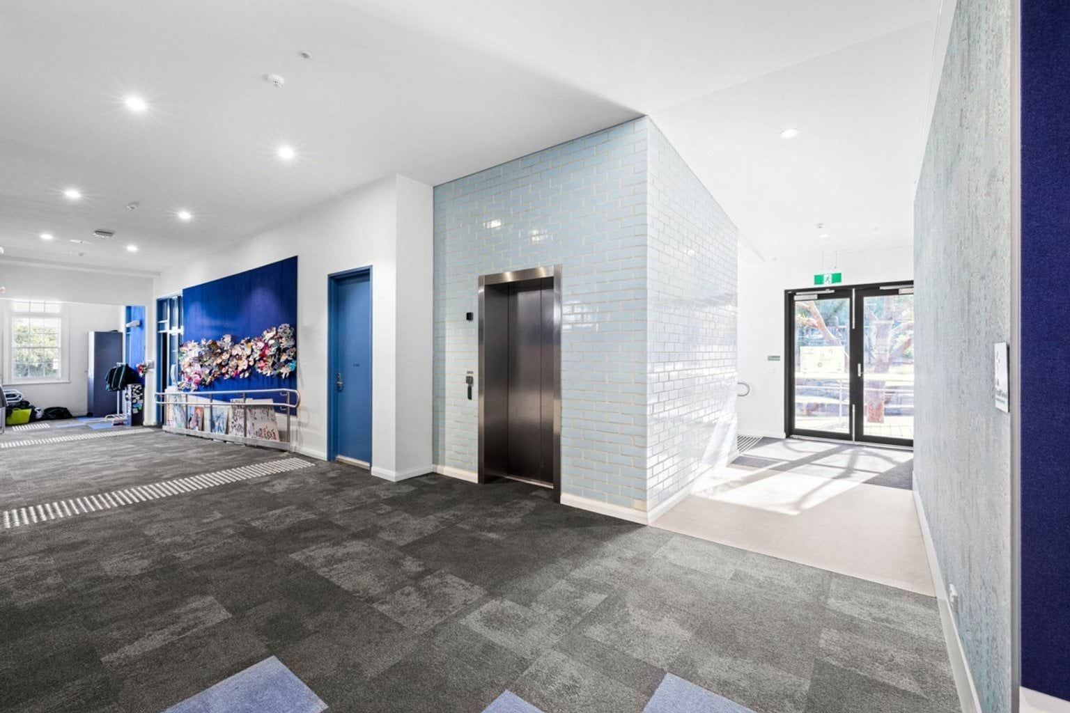 A school lobby with a stainless steel lift, blue tiled walls, and a bright entrance with glass doors. A mural featuring colourful flowers decorates one wall.