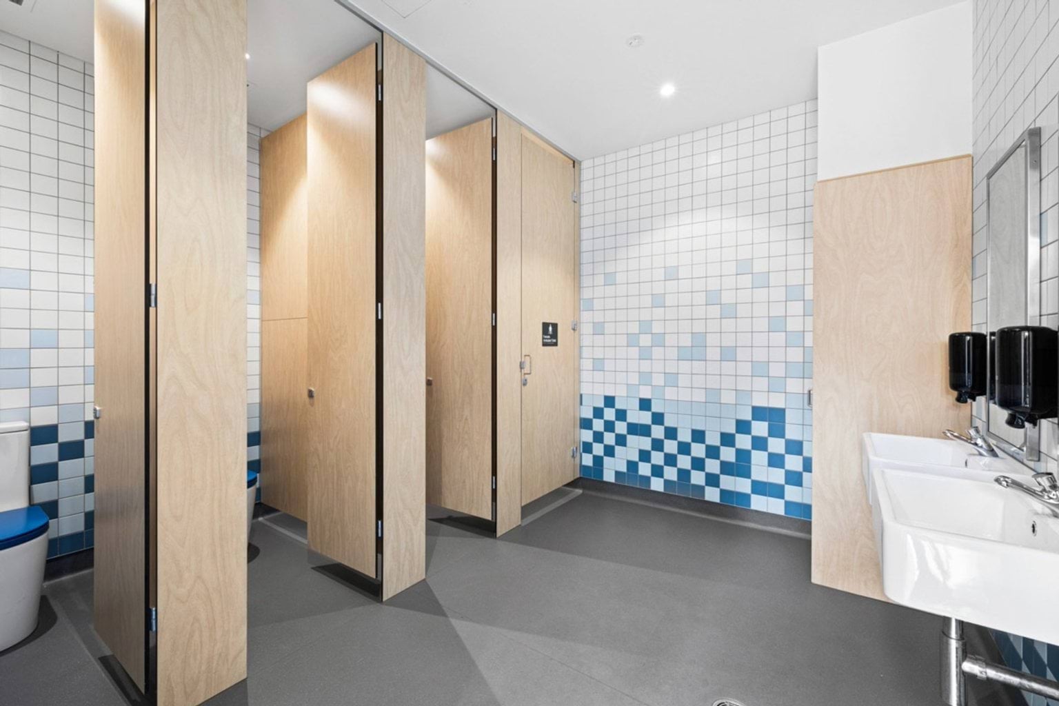 A modern student bathroom with timber cubicle doors, white and blue tiled walls, and two sinks with soap dispensers.