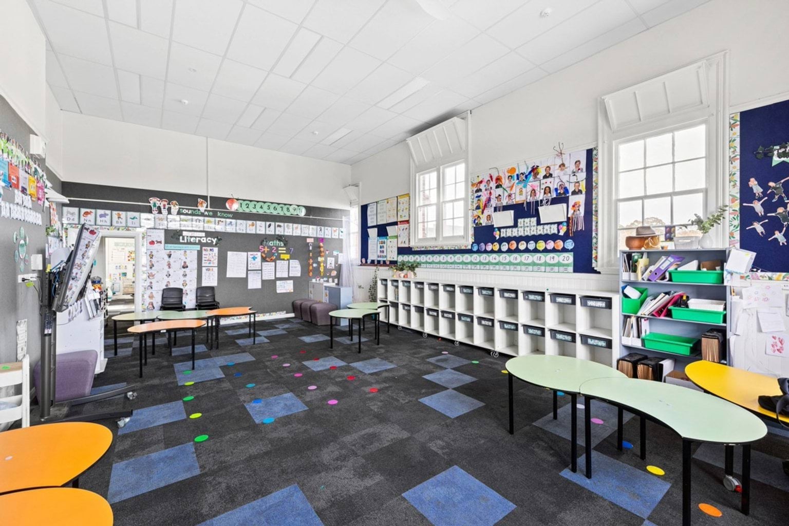 A classroom with curved tables, colourful chairs, and blue and grey carpet tiles. Student work and learning resources are displayed on the walls.