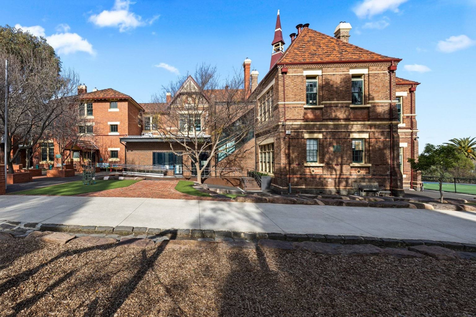 A heritage red brick school building with pitched roofs and chimneys, surrounded by landscaped paths and garden beds.