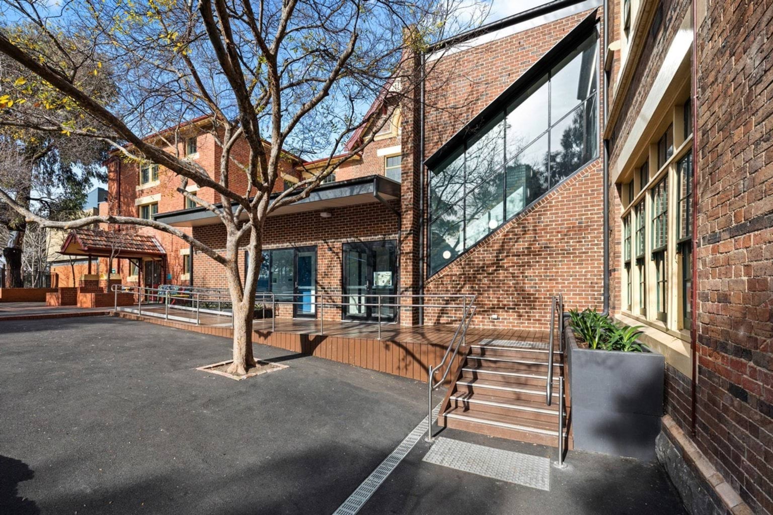 A courtyard with an accessible ramp and stairs leading to a modern glass extension on a heritage red brick school building.