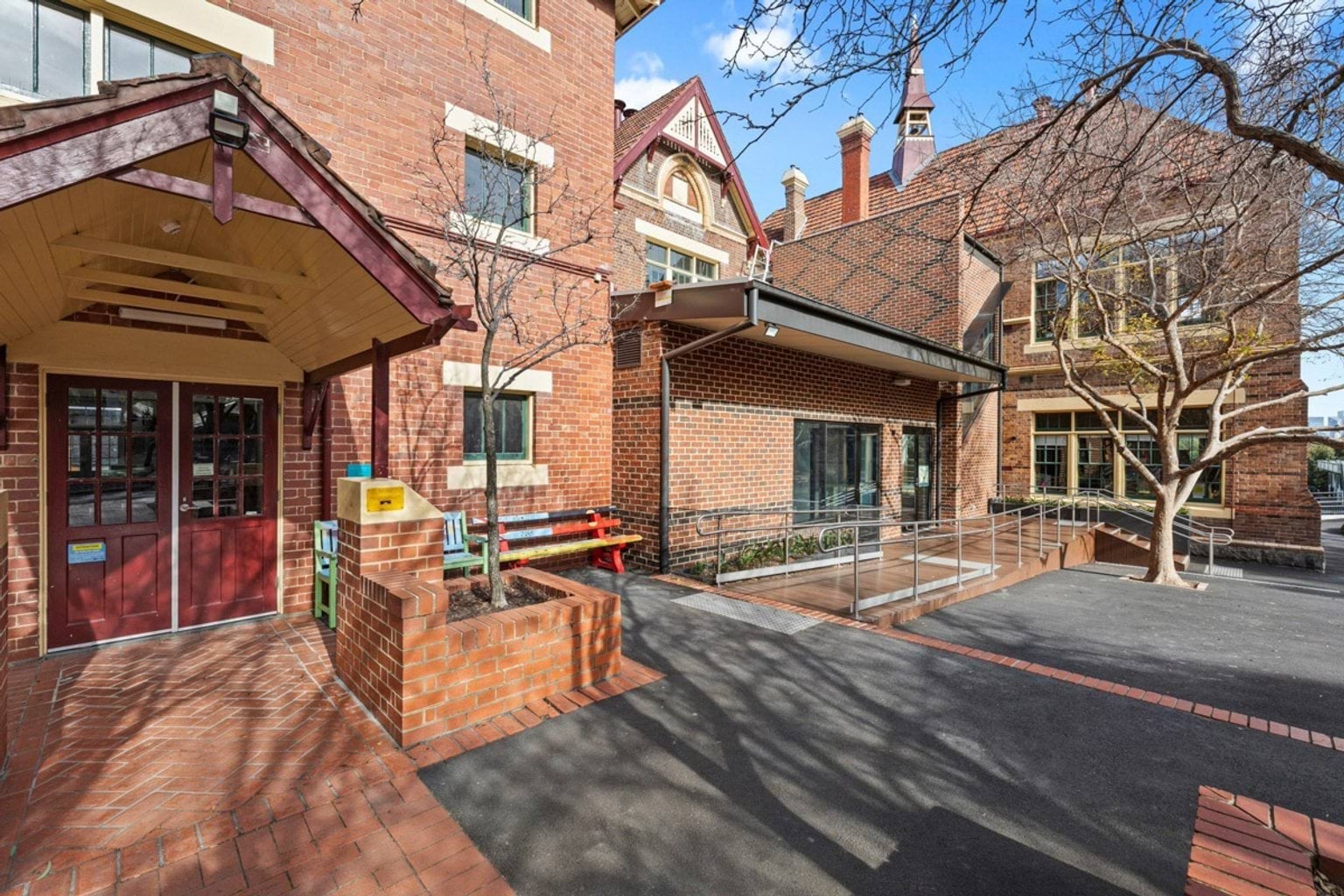 The entrance to a heritage school building with red brick walls, a covered doorway, and an accessible ramp leading to glass doors.