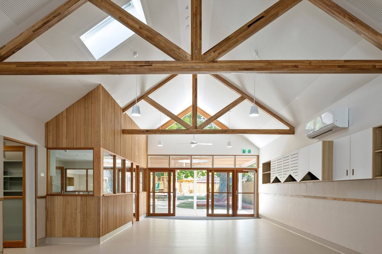 The interior of the kindergarten with a pitched ceiling and exposed timber trusses. Timber-framed glass doors open to the outdoor area, and built-in storage runs along one wall.