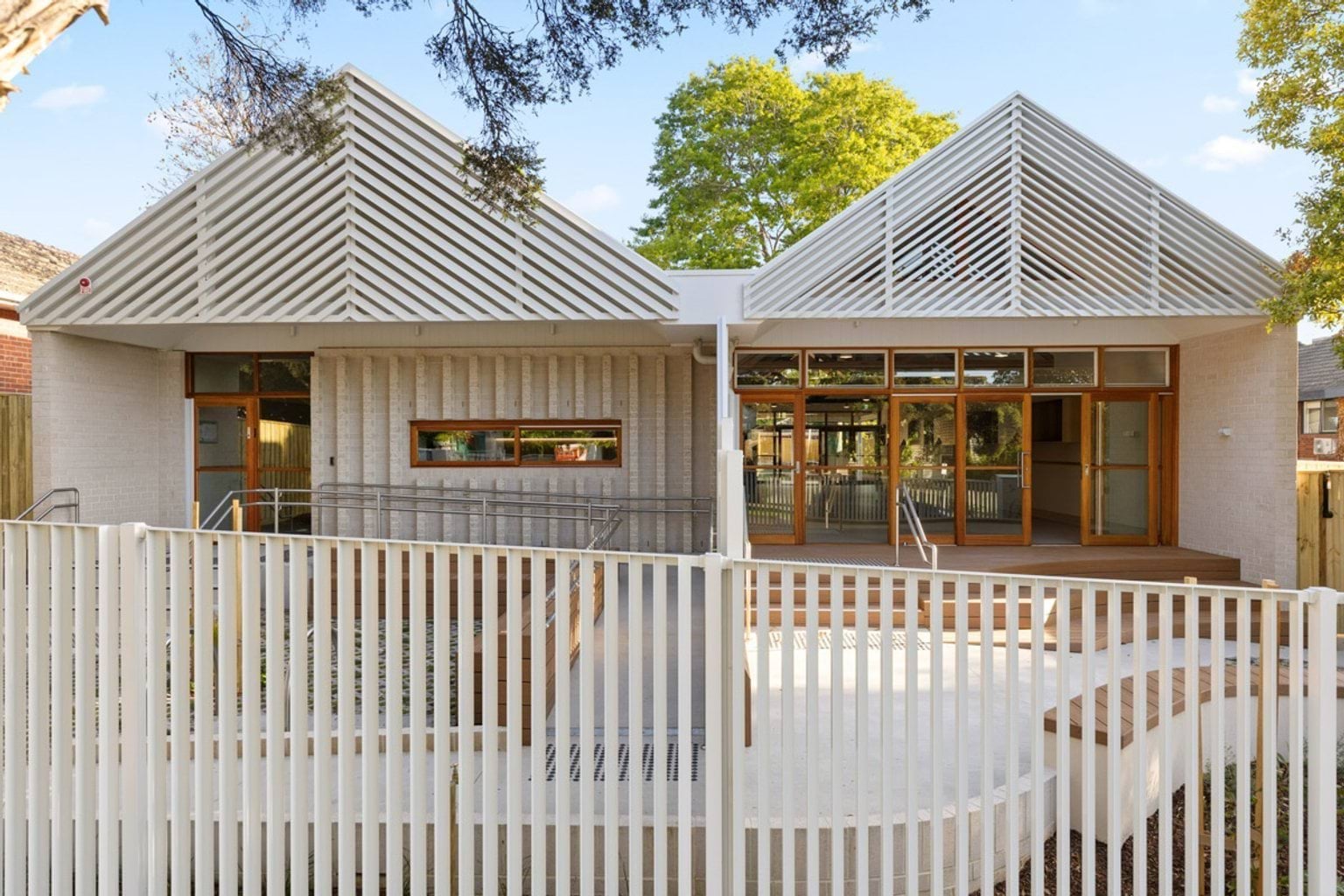 The exterior of the kindergarten with two pitched roofs featuring white slatted screens. Timber-framed glass doors open onto a raised deck, and a white fence runs along the front.