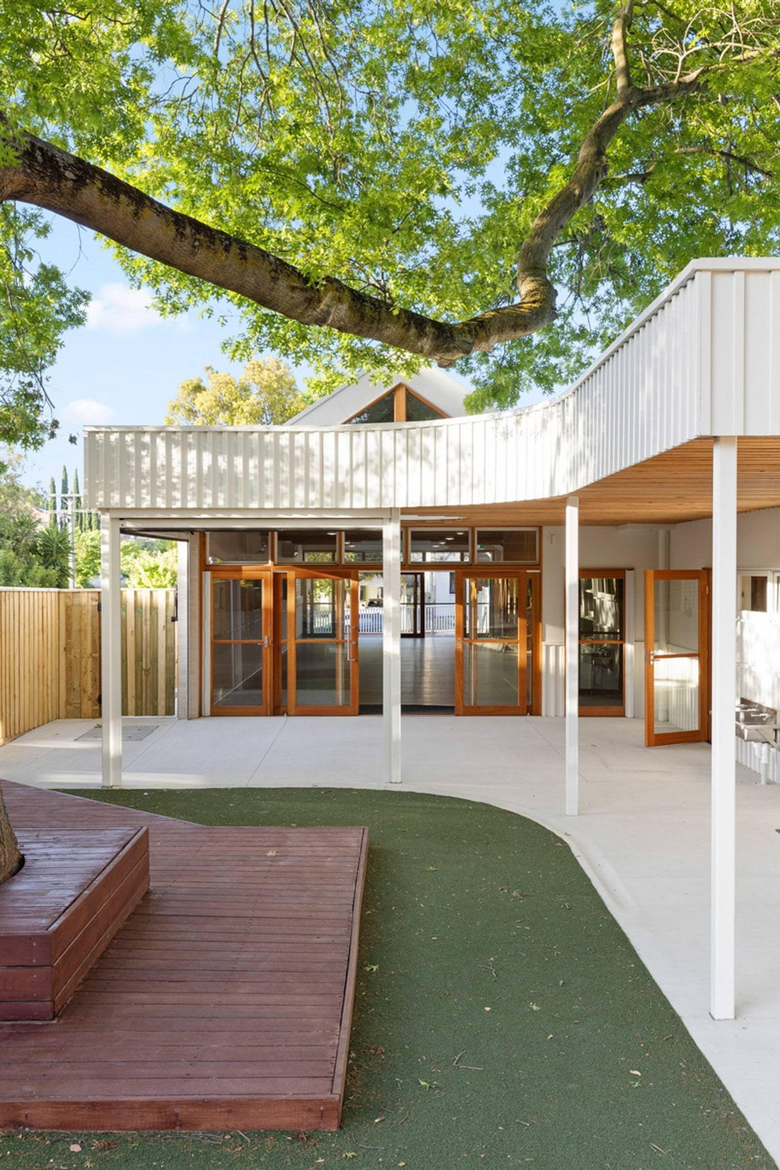 The exterior of the kindergarten with white walls, timber-framed glass doors and a curved veranda roof. A large tree provides shade over a timber deck and artificial turf area.