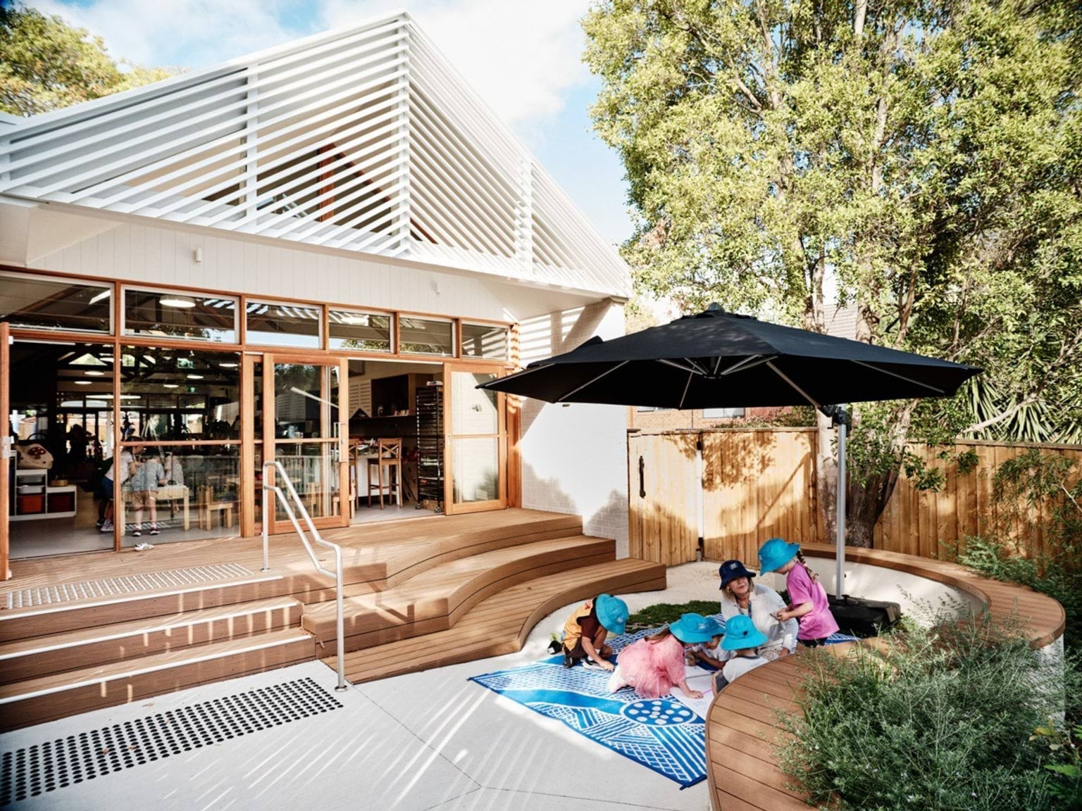 An outdoor play area with timber steps leading to glass doors. A curved seating wall surrounds a garden bed, and a large black umbrella shades a blue picnic rug.
