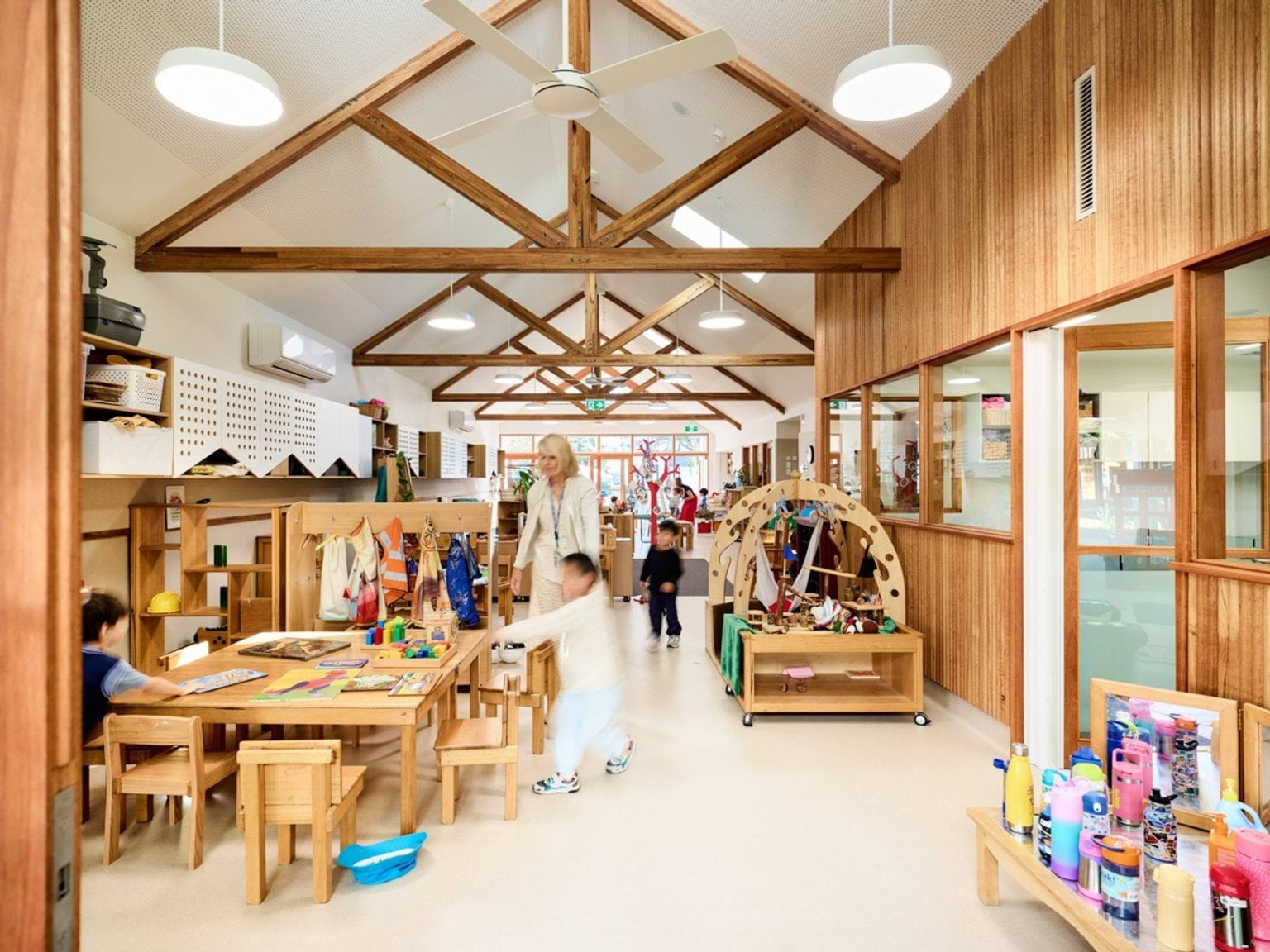 A kindergarten classroom with timber furniture and play equipment arranged along the walls. The pitched ceiling has exposed timber trusses and ceiling fans, and natural light streams through large windows.