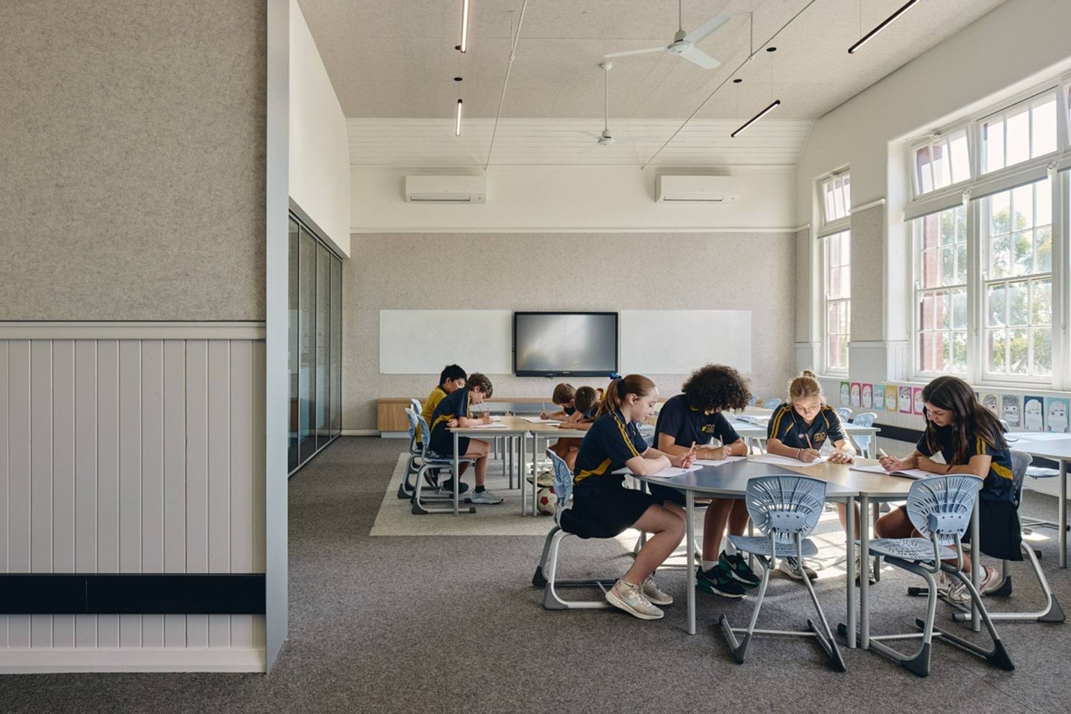 Classroom with students seated at round tables writing, large windows providing natural light and interactive screen at front