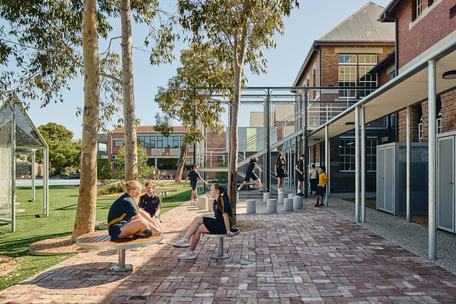 Outdoor learning space with brick paving, seating and shade trees, students sitting and talking near school buildings
