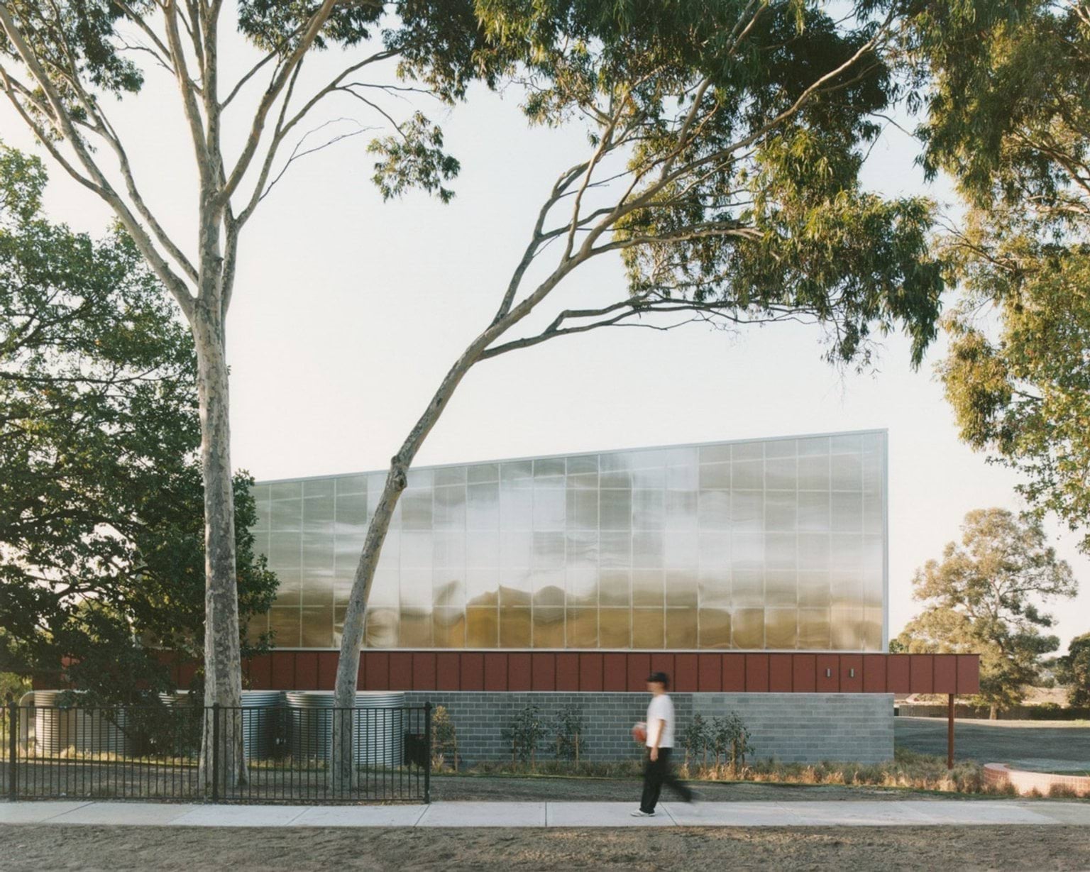 A side view of a school building with translucent wall panels and red cladding above grey brickwork. A large tree frames the image, and a person walks along the footpath outside the fence.