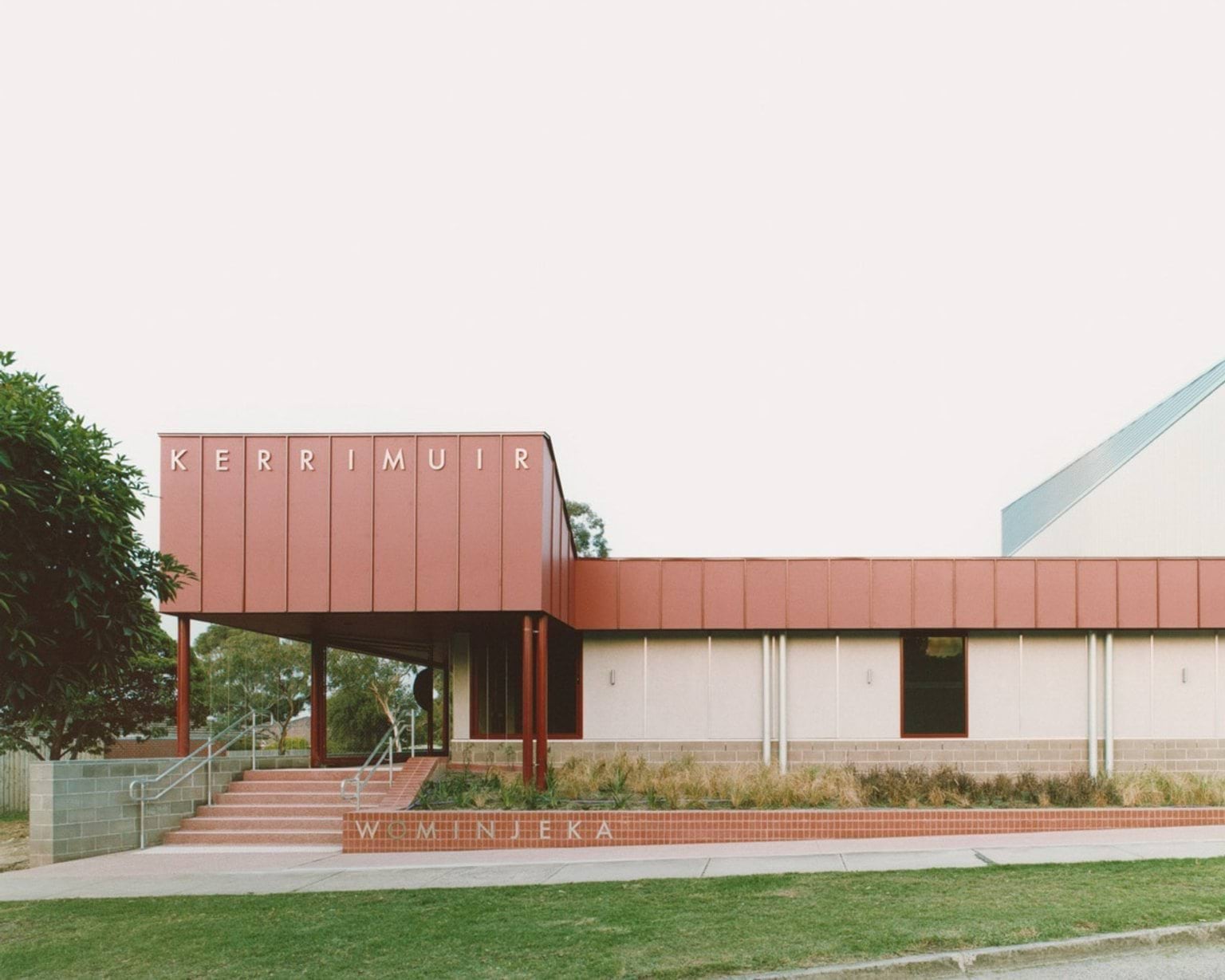 The front view of the school entrance with bold red cladding and “KERRIMUIR” signage above. Steps and landscaped garden beds lead to the doorway.