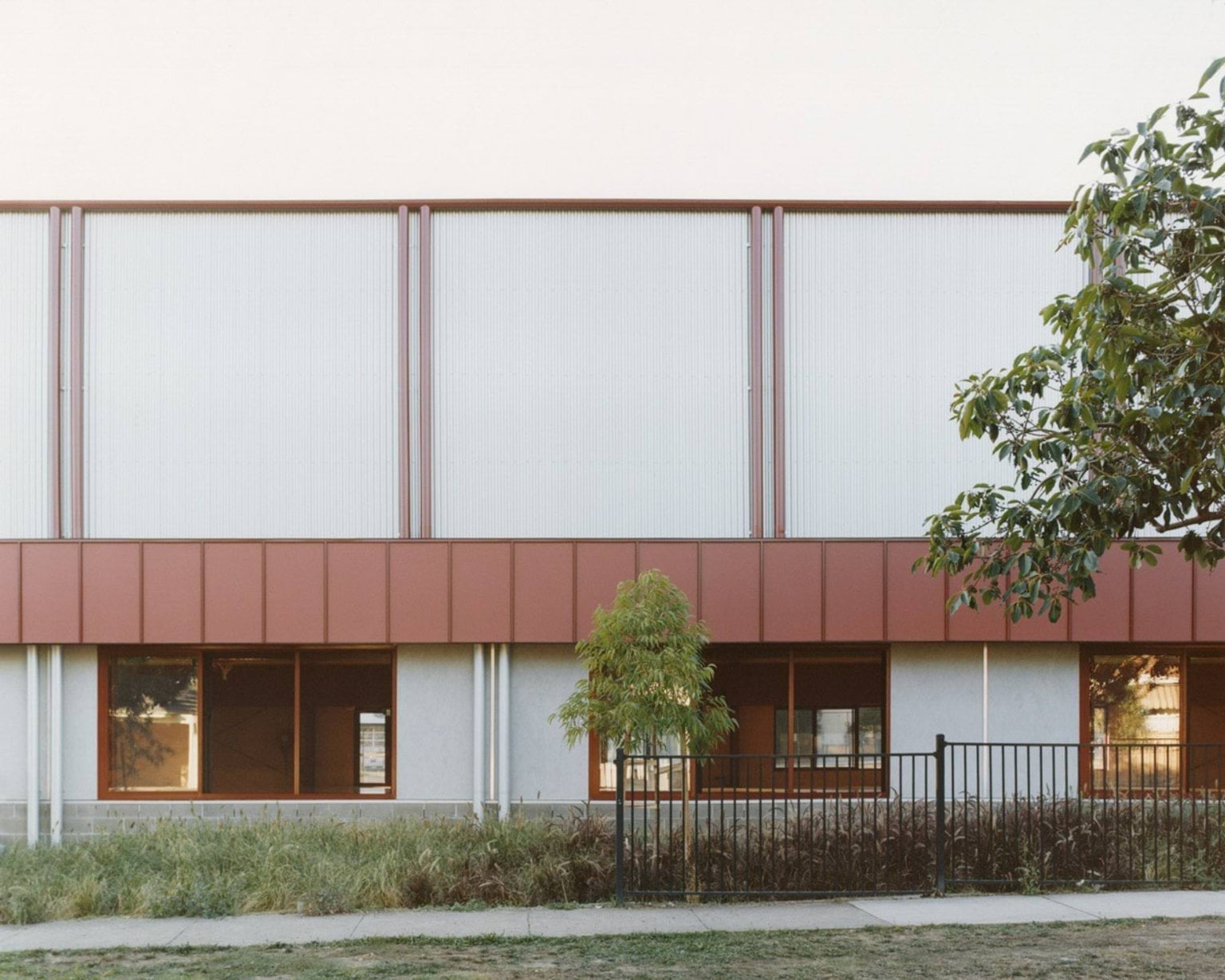 A side elevation of the school building with red cladding and translucent panels above grey walls. A black metal fence and young trees line the footpath.