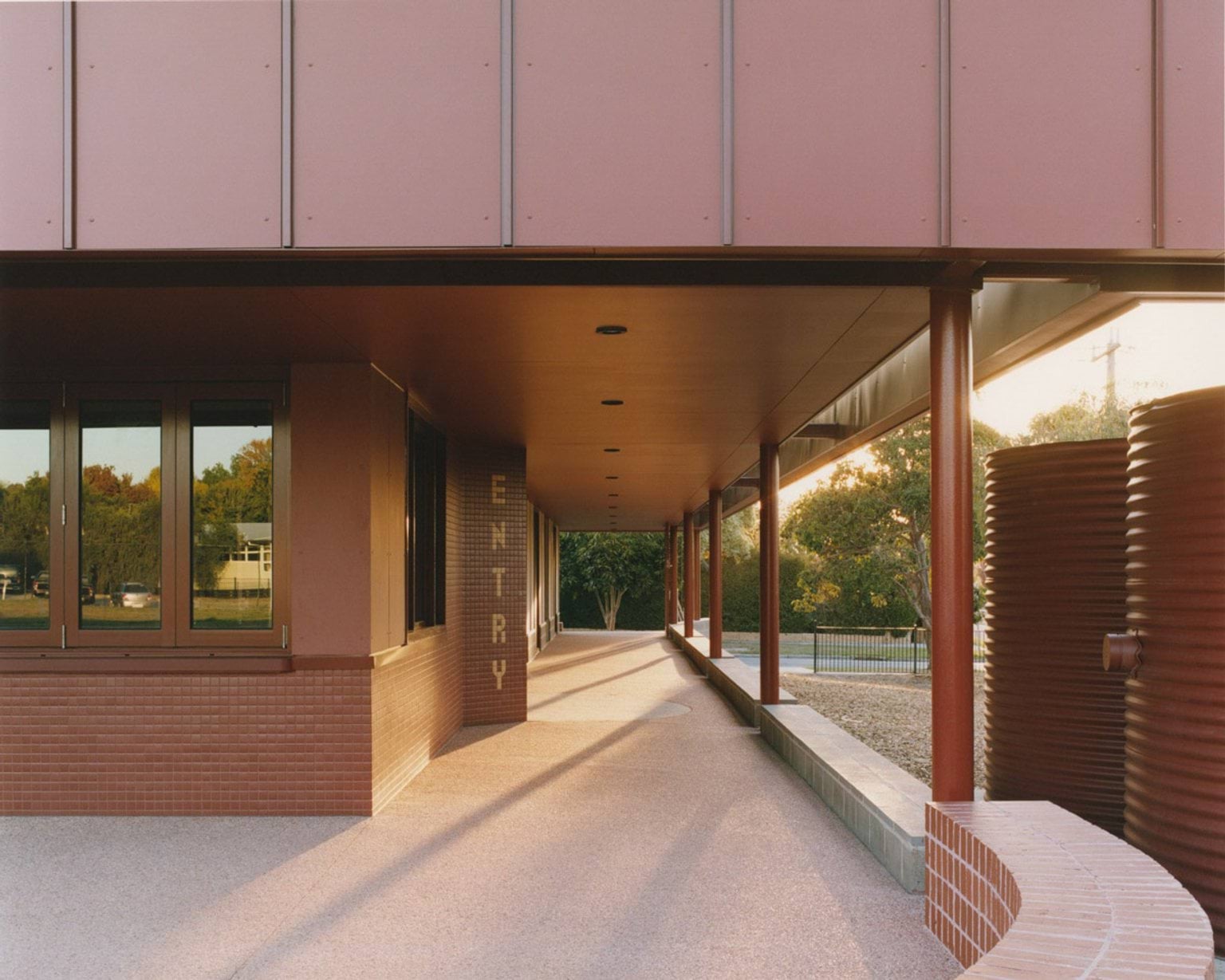 A covered walkway with red cladding and brick walls leading to the school entry. Large cylindrical water tanks are visible to the right.