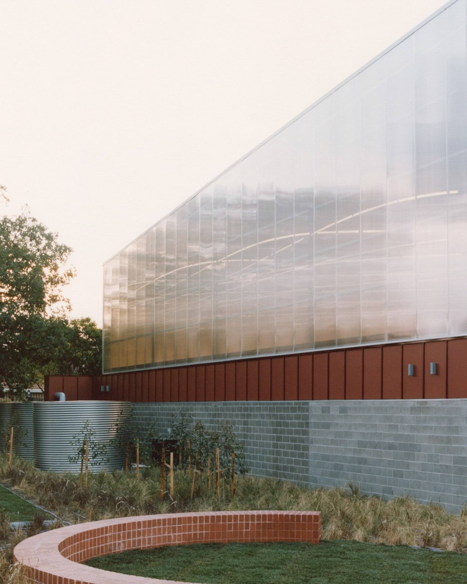 The exterior of a school building with translucent wall panels and red cladding above grey brickwork. A curved brick seating wall and landscaped garden are in the foreground.