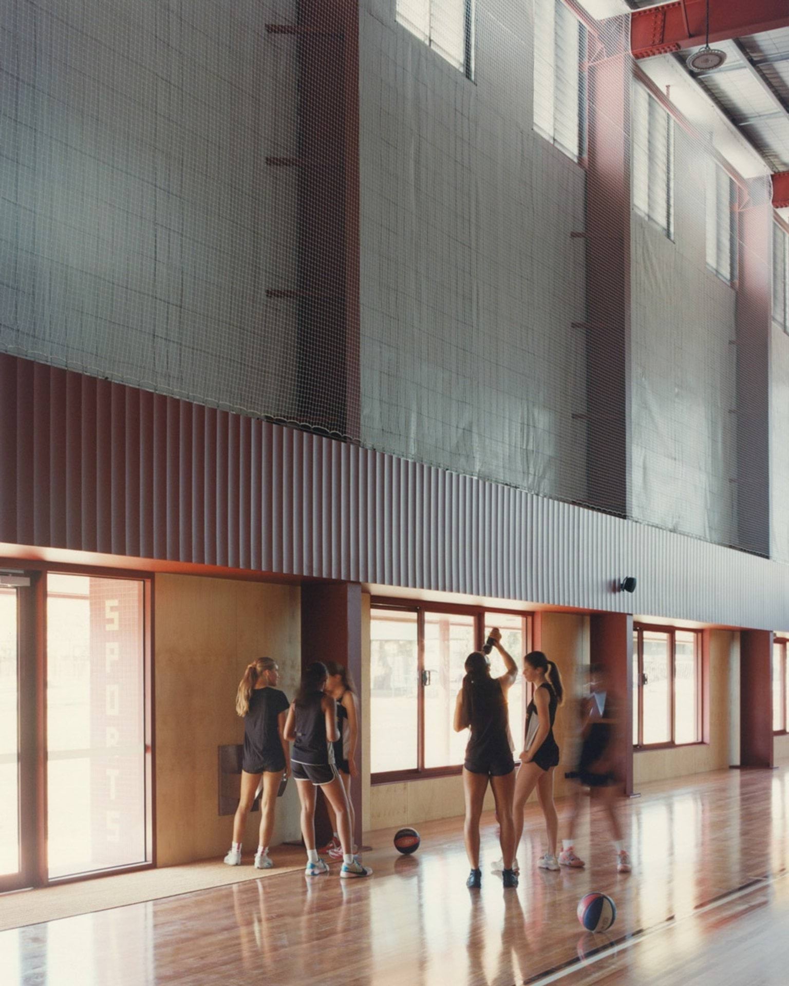 An indoor sports court with timber flooring and high ceilings. A group of students stands near the wall with basketballs on the floor.