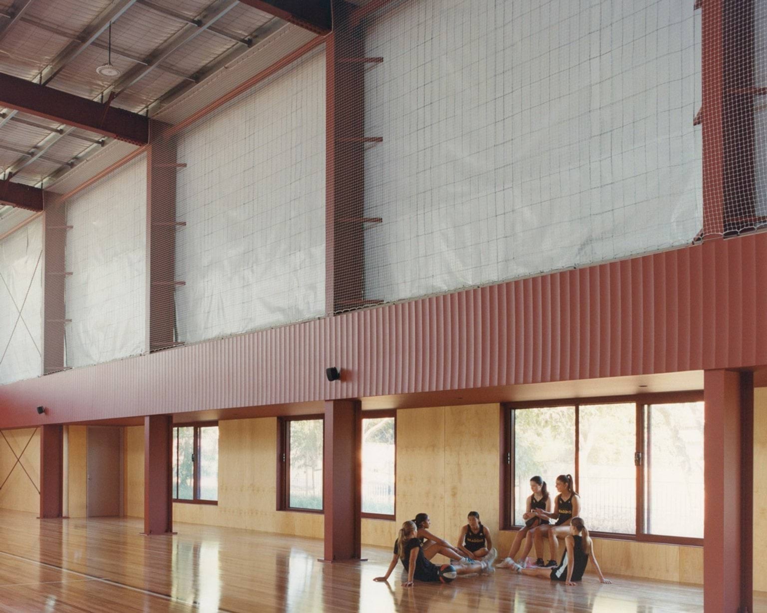 An indoor sports court with timber flooring and high ceilings. A group of students stands near the wall with basketballs on the floor.