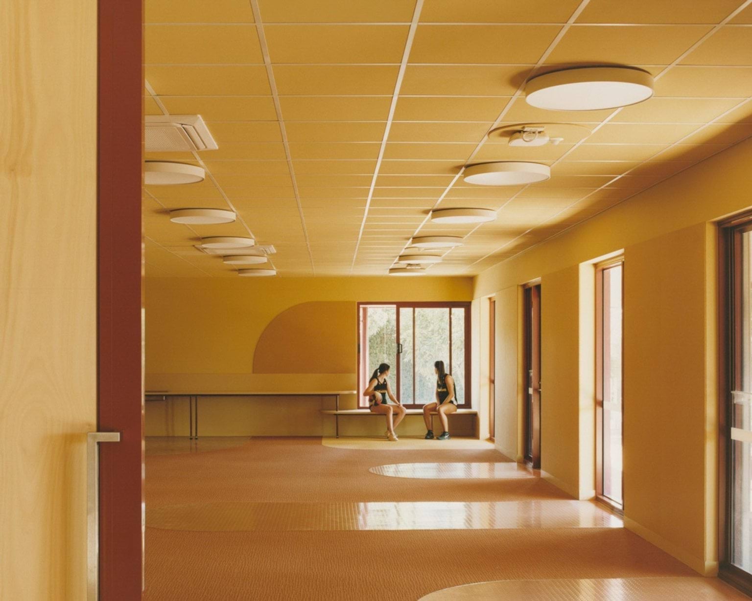 An indoor learning space with yellow walls, round ceiling lights, and timber-framed windows. Two students sit on a bench near the window.