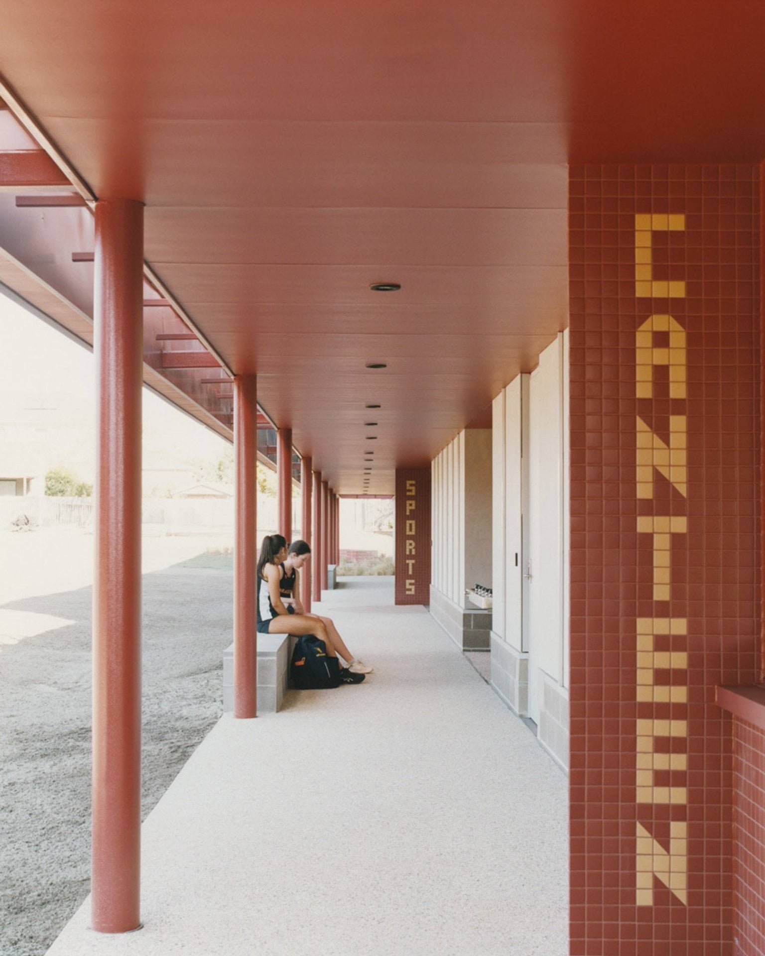 A covered walkway with red cladding and tiled signage reading “CANTEEN.” Two students sit on a bench under the walkway.