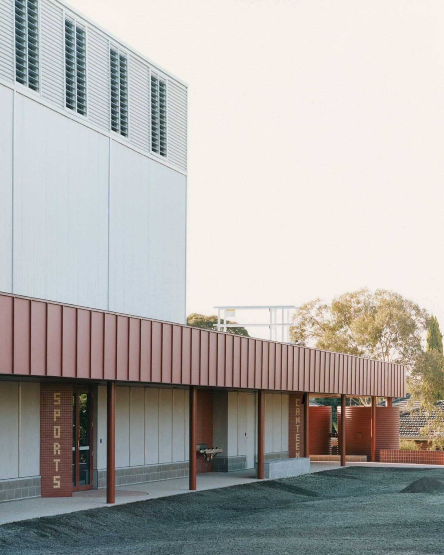 A school building with red walls and signage for “SPORTS” and “CANTEEN.” A covered walkway runs along the building, and water tanks are visible in the background.