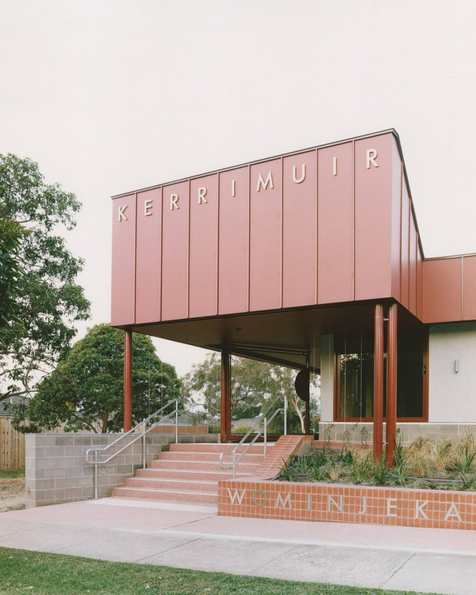 The main entrance of the school with bold red cladding and the word “KERRIMUIR” displayed above. Wide steps lead to the doorway, and “WOMINJEKA” is tiled on the lower step.