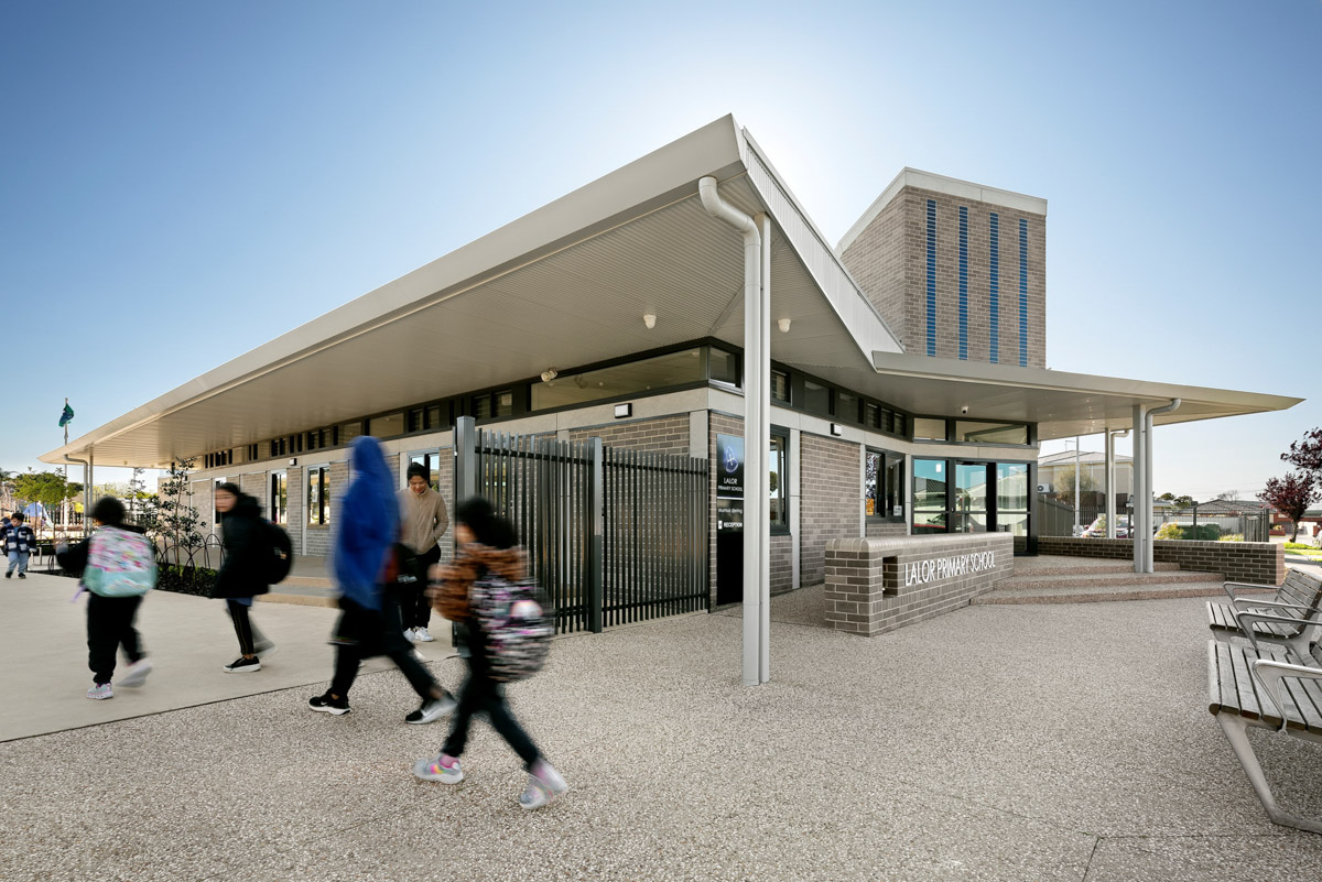 The main entrance of a modern school building with grey brick walls, angled rooflines, and a sign reading “Lalor Primary School.” Students are walking outside.