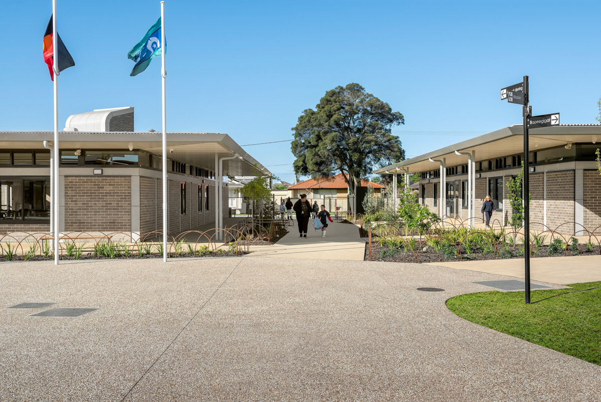 A landscaped courtyard with two flagpoles, garden beds, and paved walkways between modern brick school buildings.