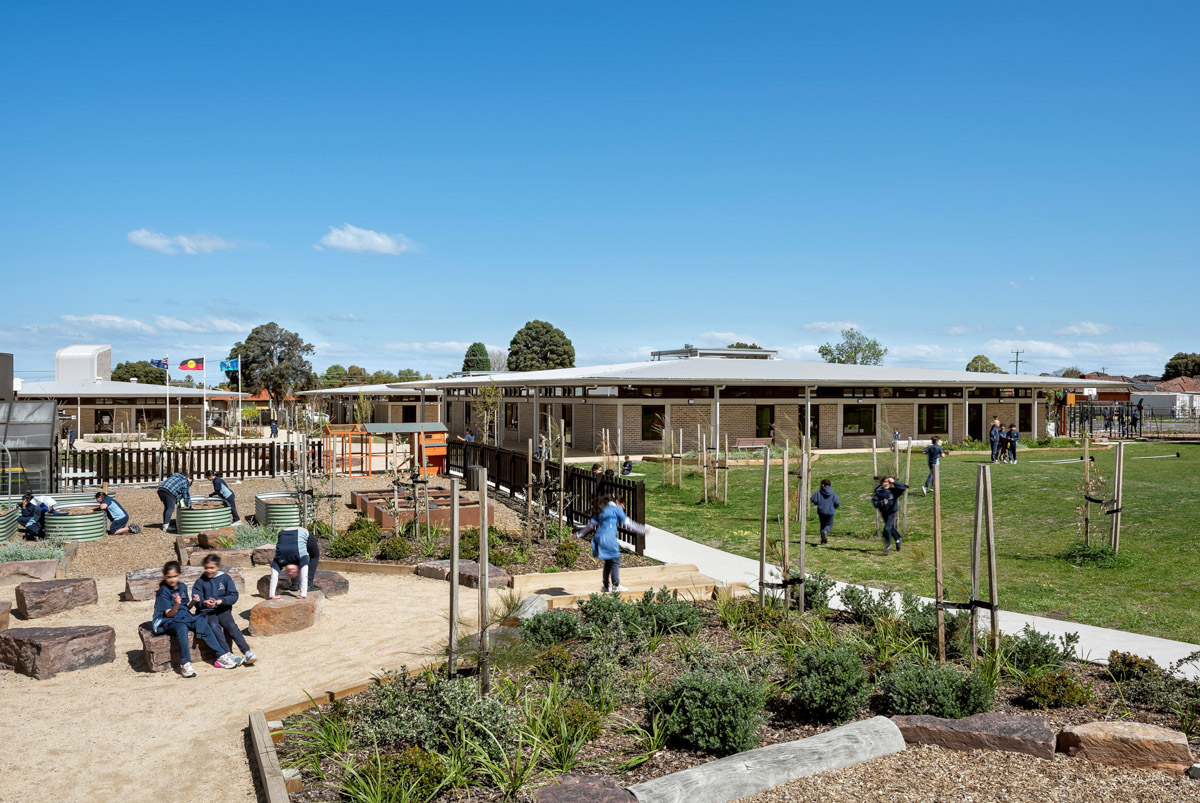 An outdoor play and garden area with raised garden beds, rocks, and young trees. Students are seated and playing under a clear blue sky.