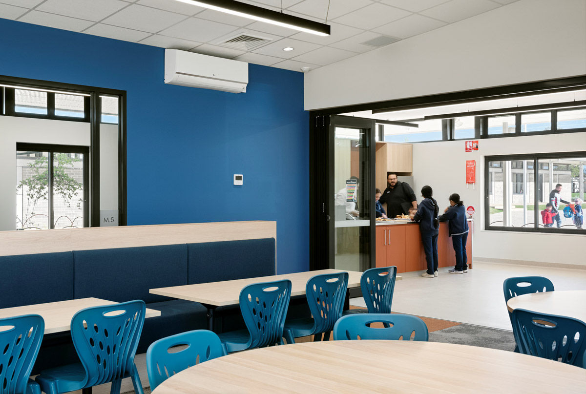 An indoor canteen space with blue walls, round tables, and blue chairs. A serving counter opens to the outdoor area through glass doors.