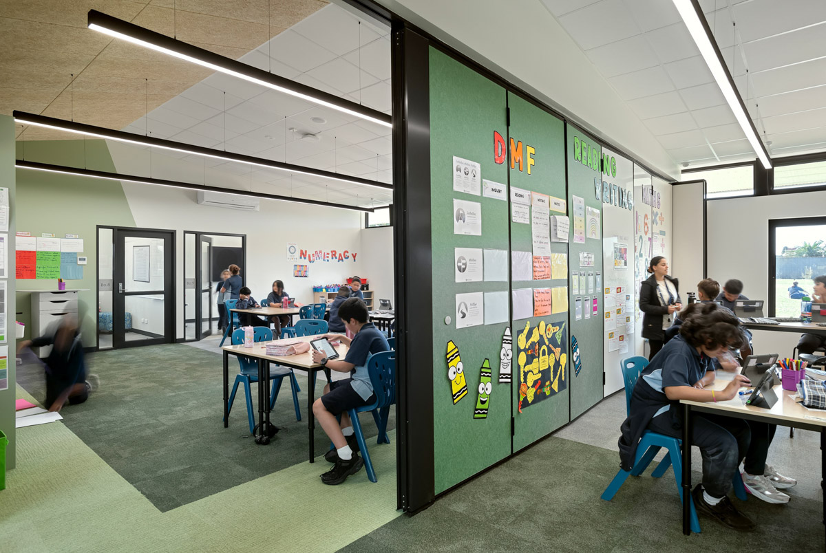 An open learning space with green carpet, movable partitions displaying student work, and tables with blue chairs arranged for group activities.