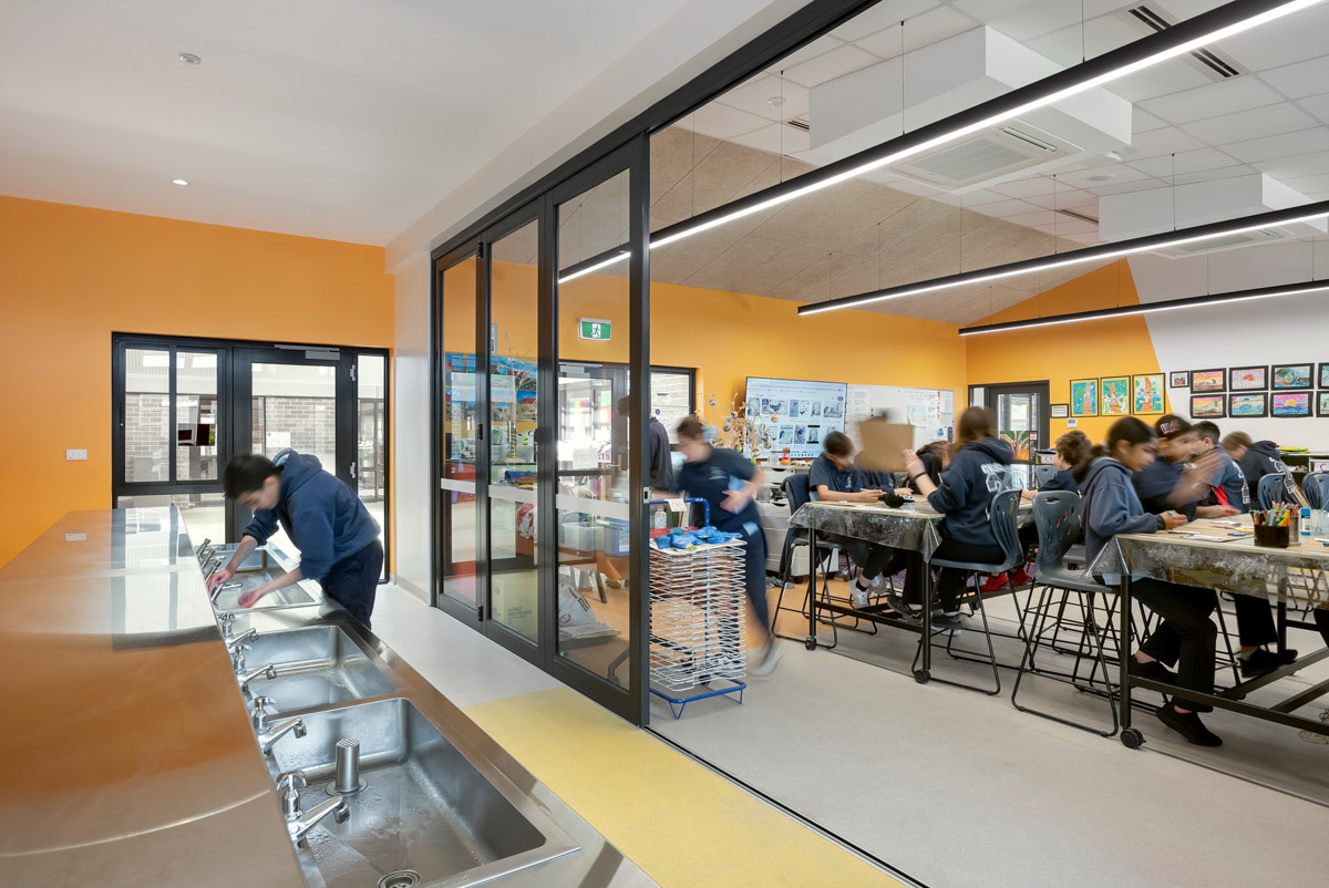 An art classroom with orange walls, stainless steel sinks along one side, and students working at tables with art supplies.