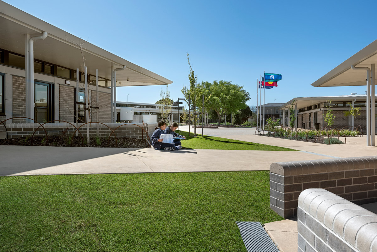 An outdoor seating area with low brick walls, garden beds, and paved paths between modern school buildings. Flags are visible in the background.