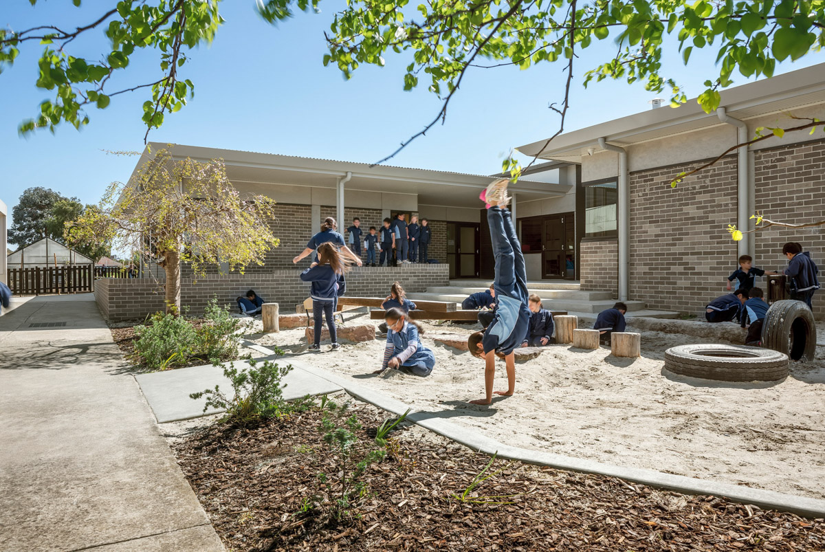 An outdoor sandpit play area with timber logs, tyres, and garden beds. Students are playing and exploring under a covered walkway.