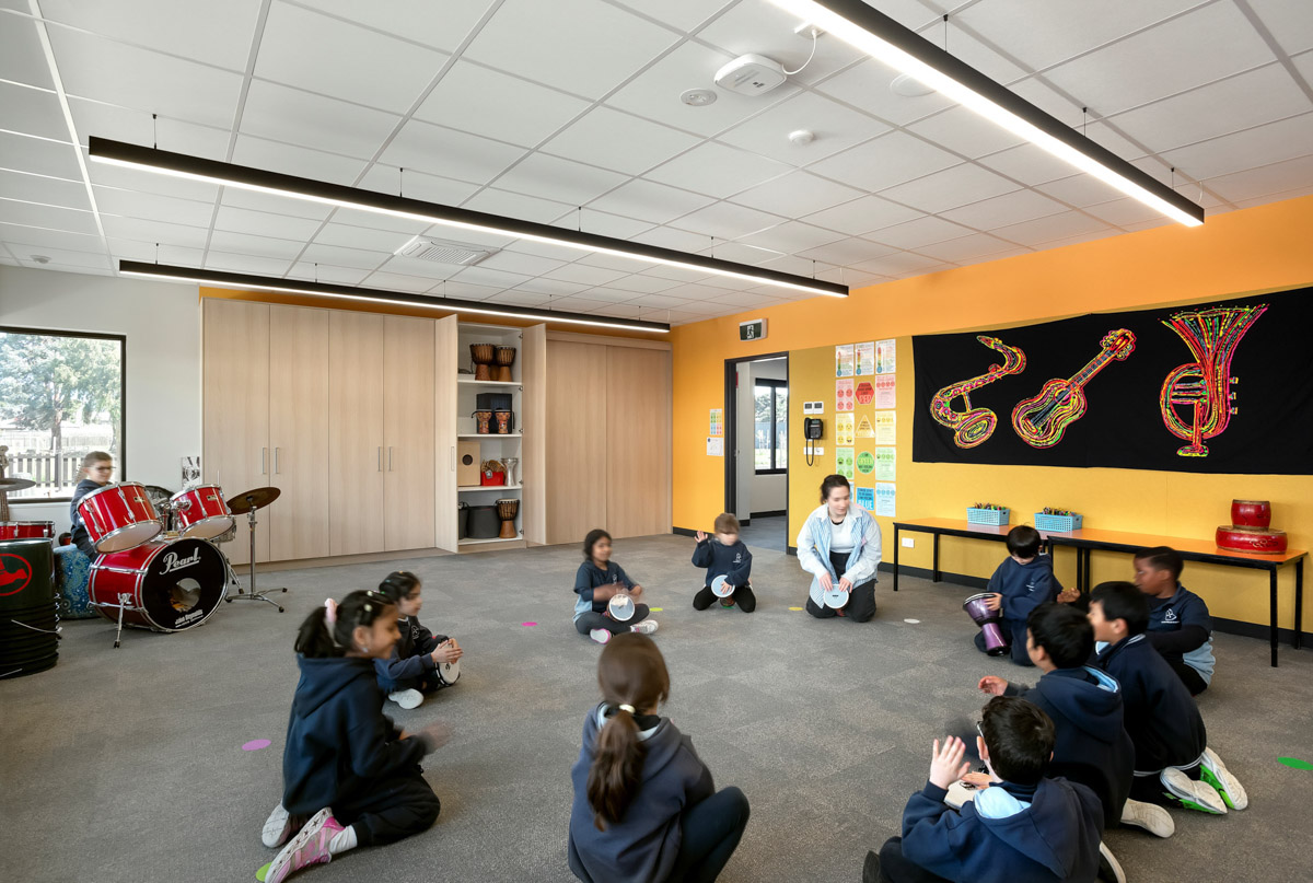 A music classroom with orange feature wall, drum kit, and percussion instruments on shelves. Students are seated in a circle on grey carpet.