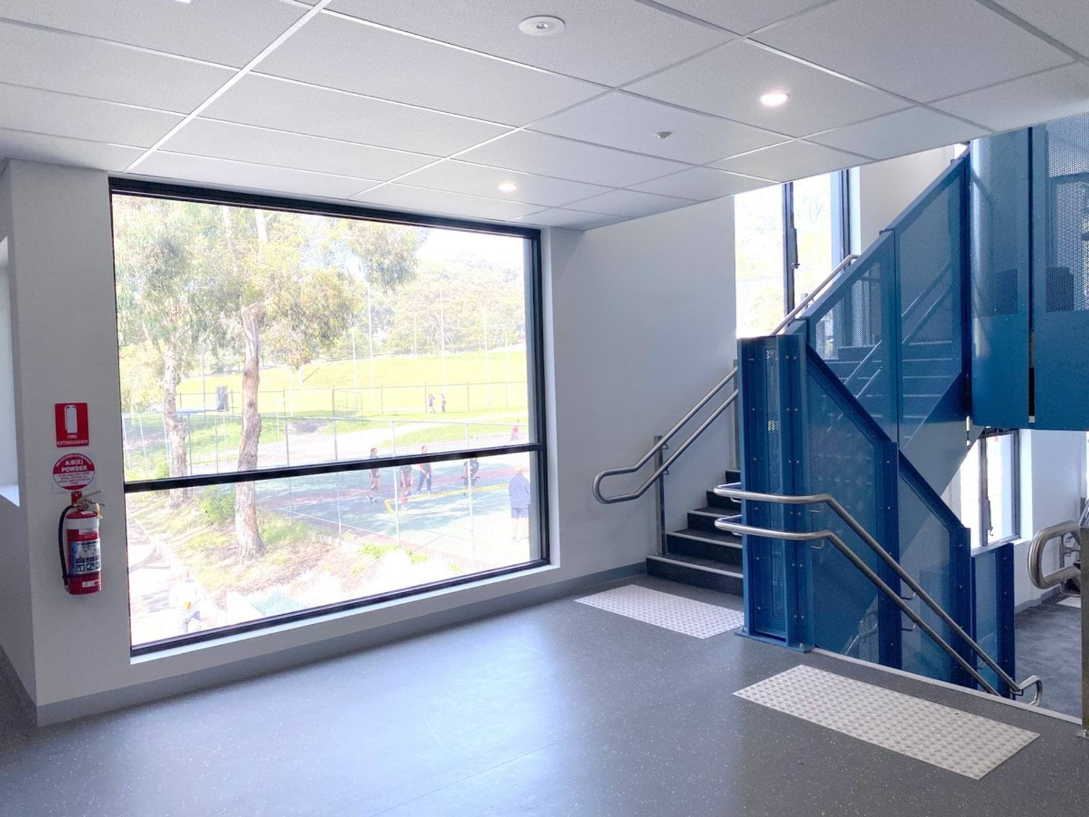 An interior staircase with blue metal panels and stainless steel handrails next to a large window overlooking outdoor sports courts.