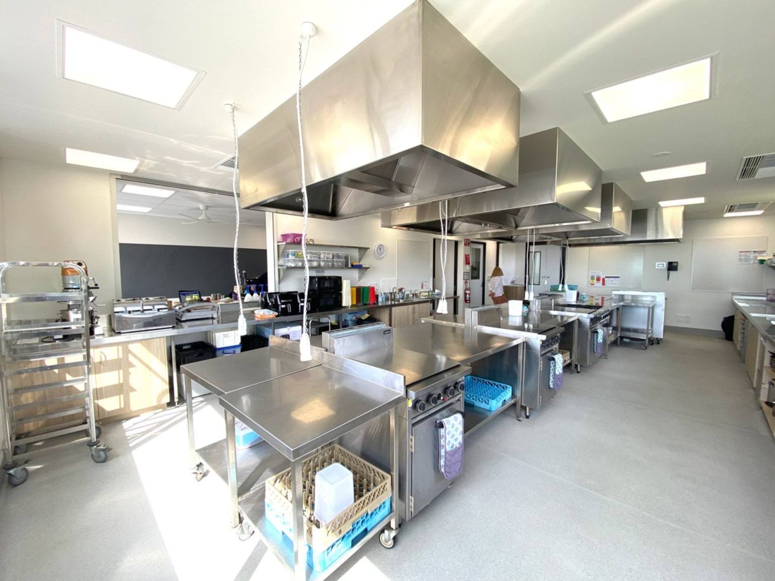 A bright commercial kitchen classroom with stainless steel benches, ovens, and large overhead range hoods. Trolleys and storage shelves line the walls.