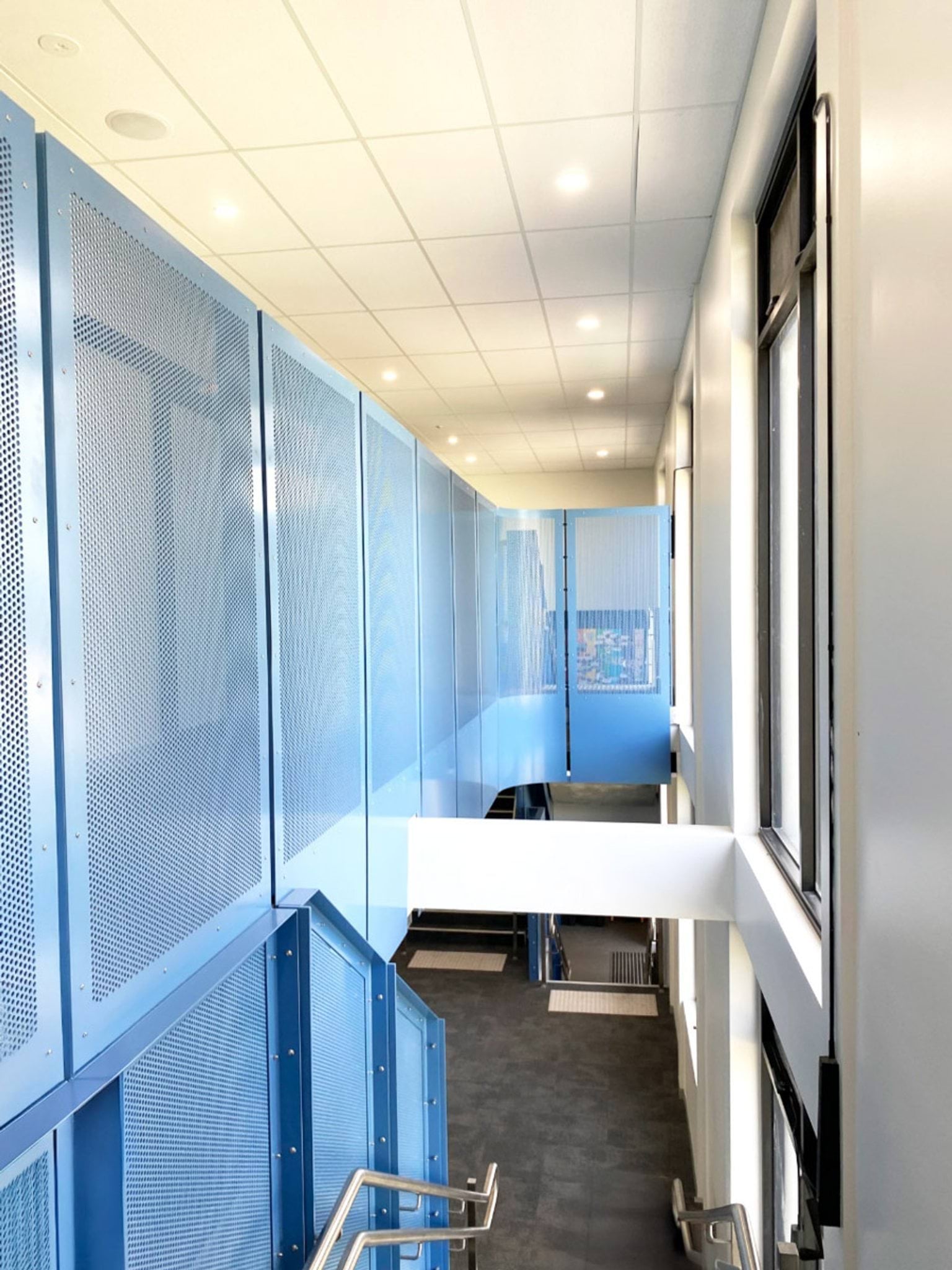 A bright hallway with grey carpet and a staircase featuring blue metal panels and stainless steel handrails. A lift is visible near the stairs.
