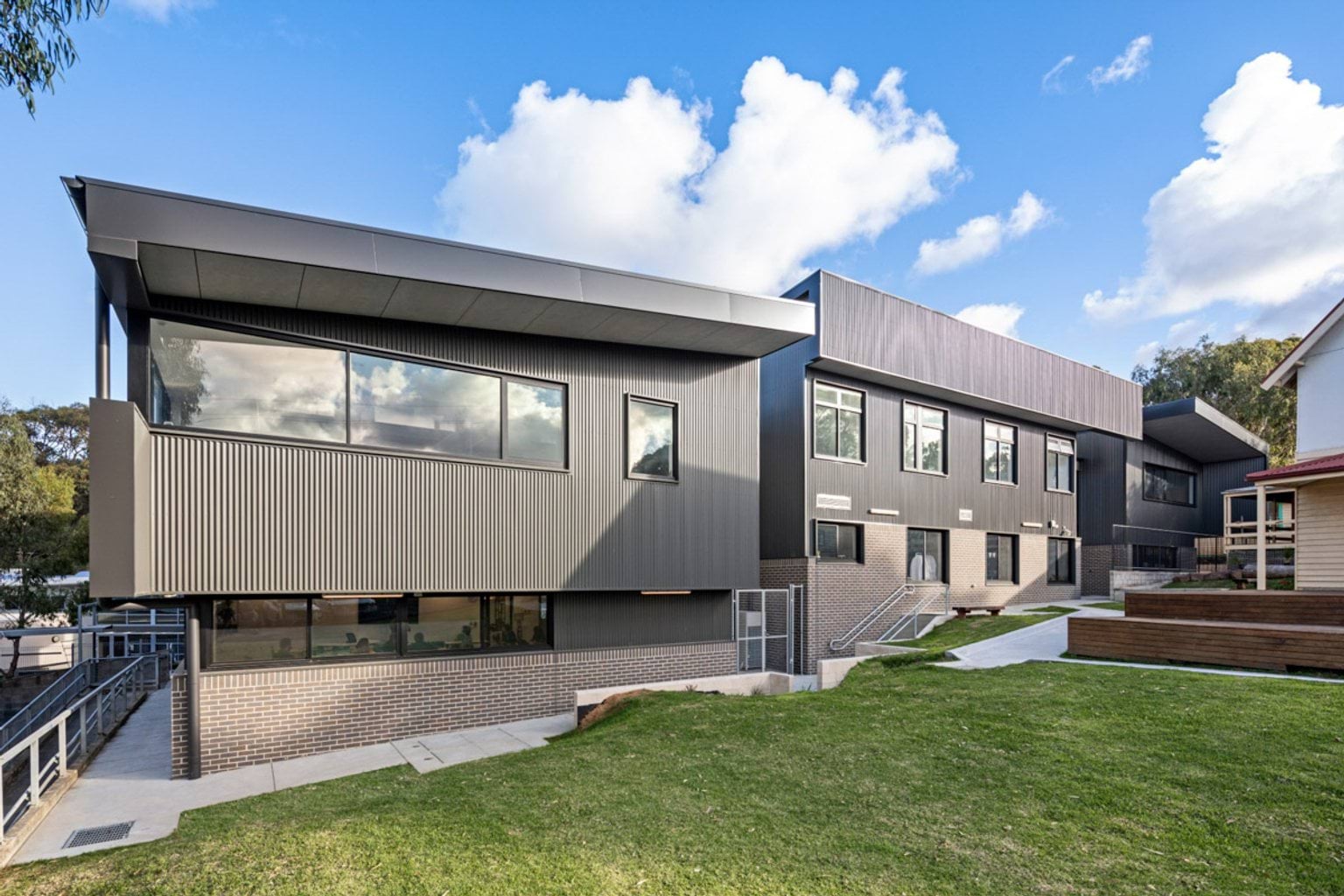 The exterior of a modern school building with grey corrugated cladding, brickwork, and large windows. A grassed area and concrete paths are in the foreground.